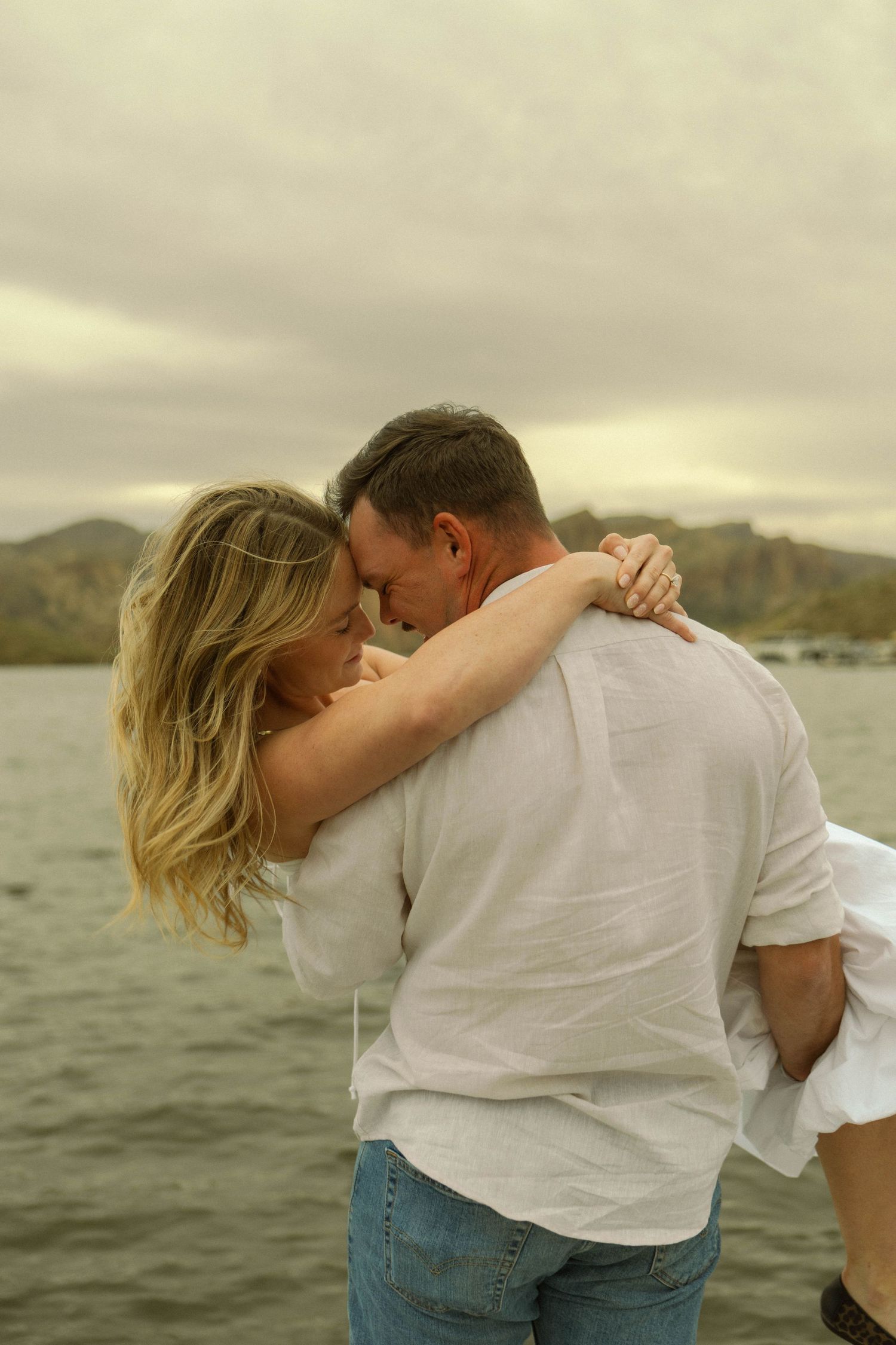 A couple in white shirts embrace romantically by a scenic lake with mountains in the background.