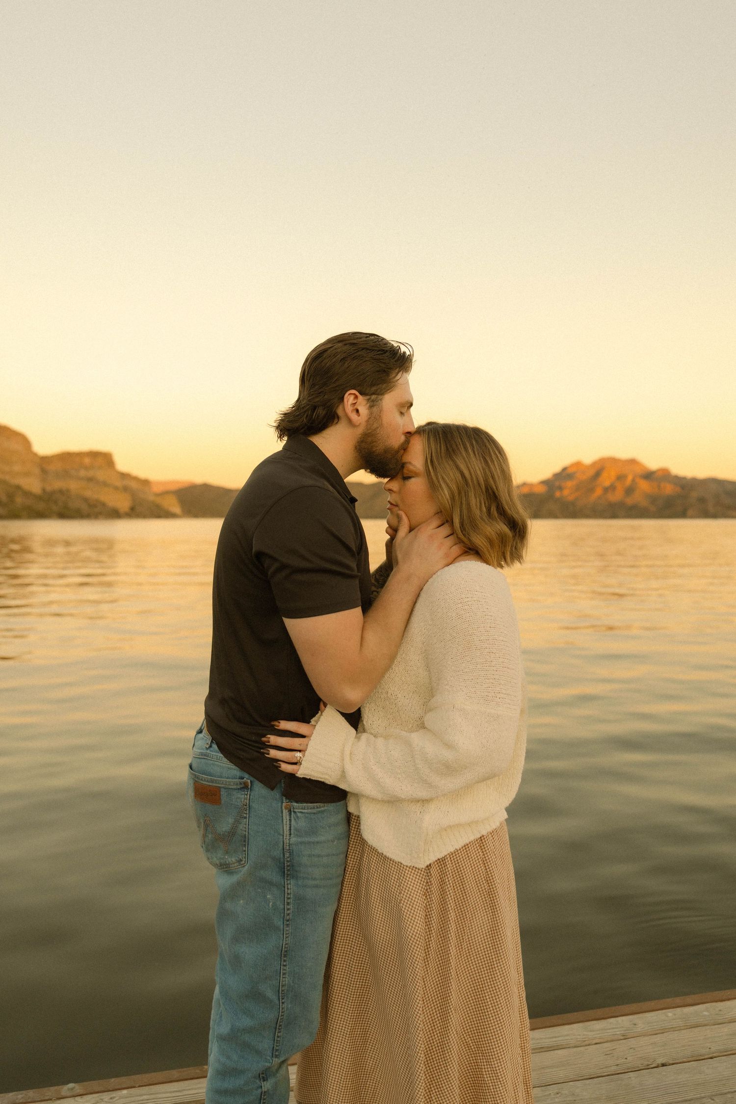 A romantic couple embraces on a dock at sunset with mountains and a lake in the background.