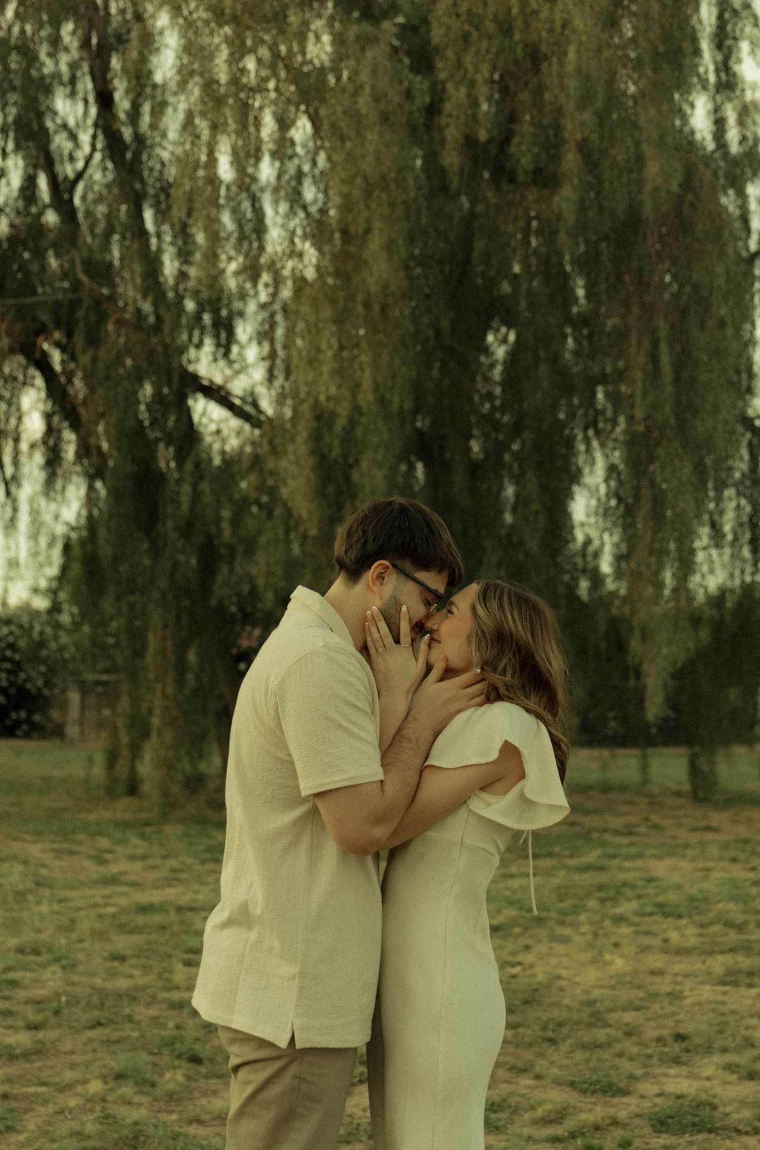A couple shares a romantic kiss outdoors beneath a weeping willow tree, both dressed in cream outfits.
