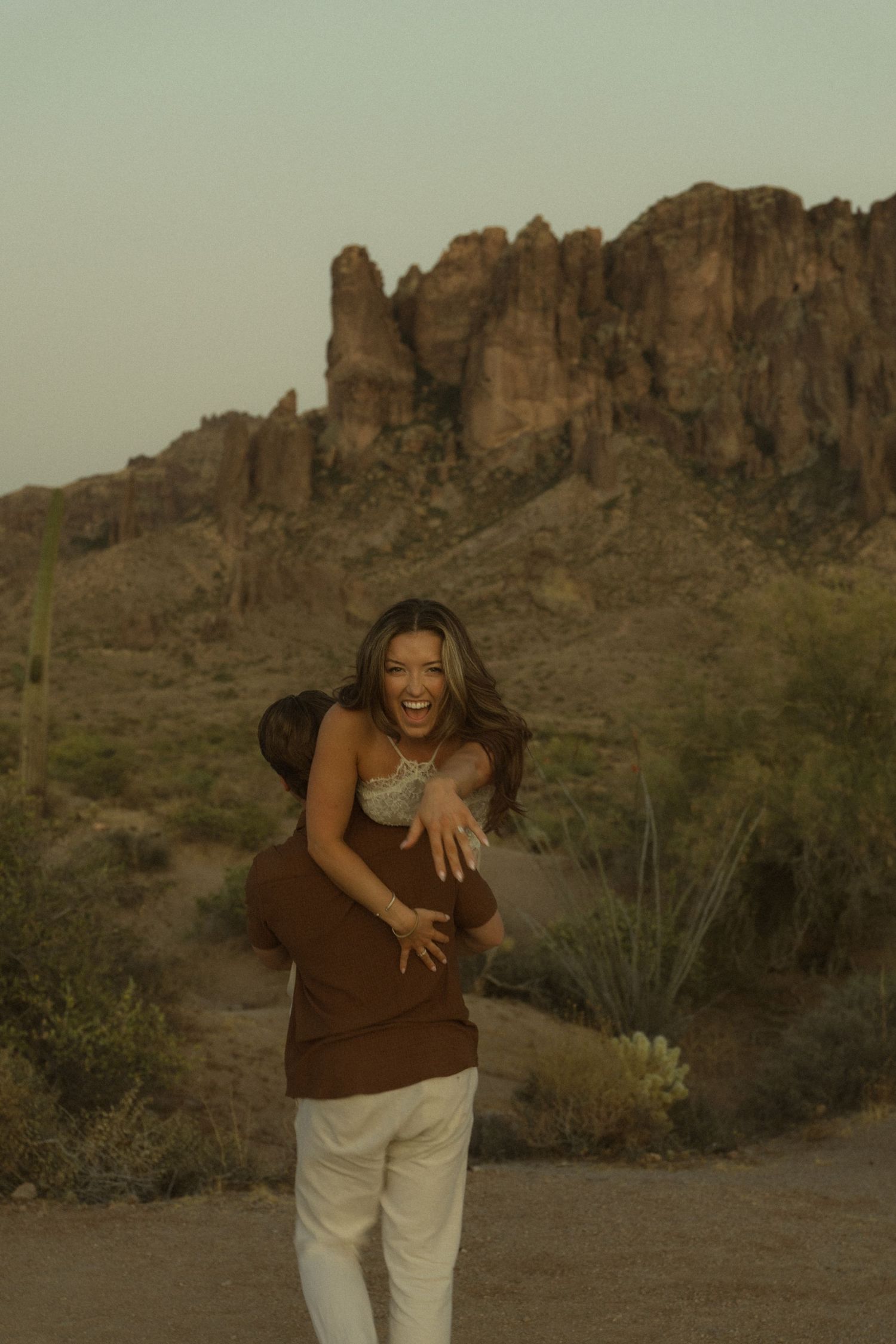 Laughing bride being lifted by groom in brown shirt with Superstition Mountains desert scenery at dusk.