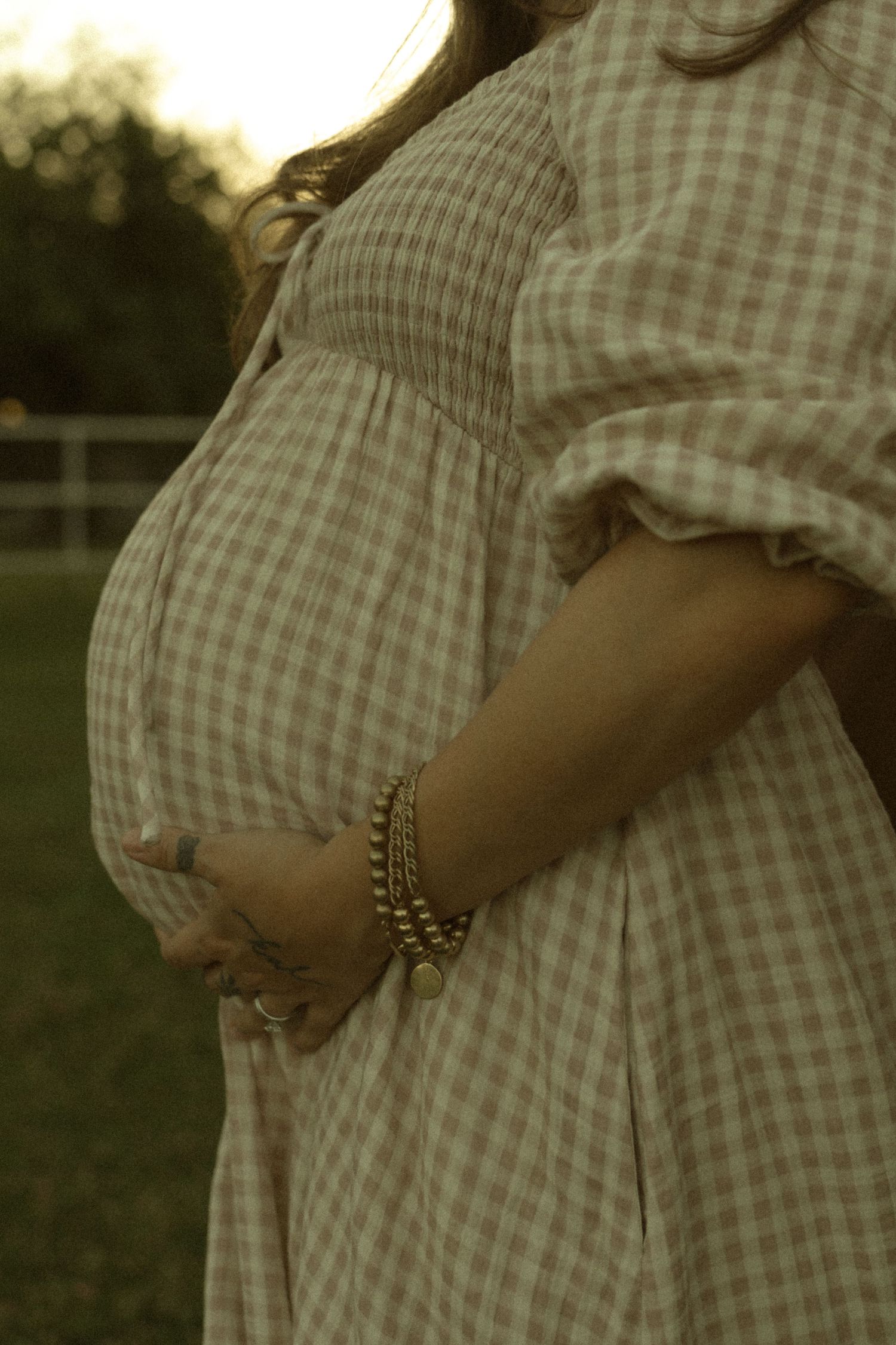 Close-up of pregnant woman in pink gingham dress cradling baby bump with gold bracelets outdoors.