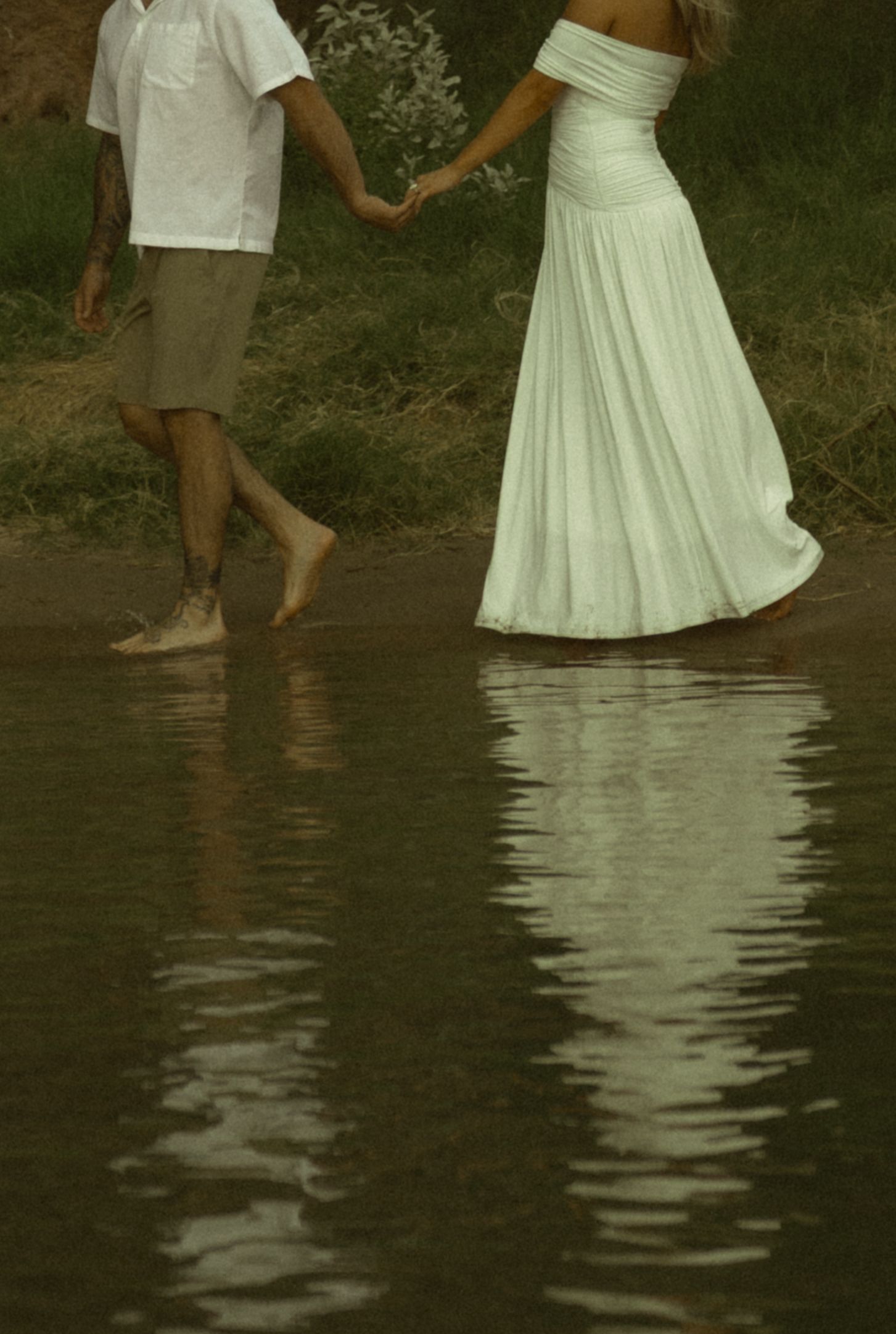 Bride in white gown and groom holding hands barefoot at the water's edge with reflections.