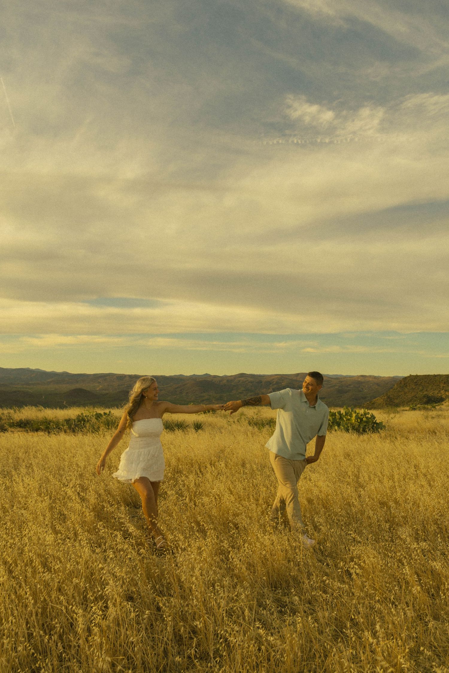 Couple playfully running through golden field at sunset with mountain views.