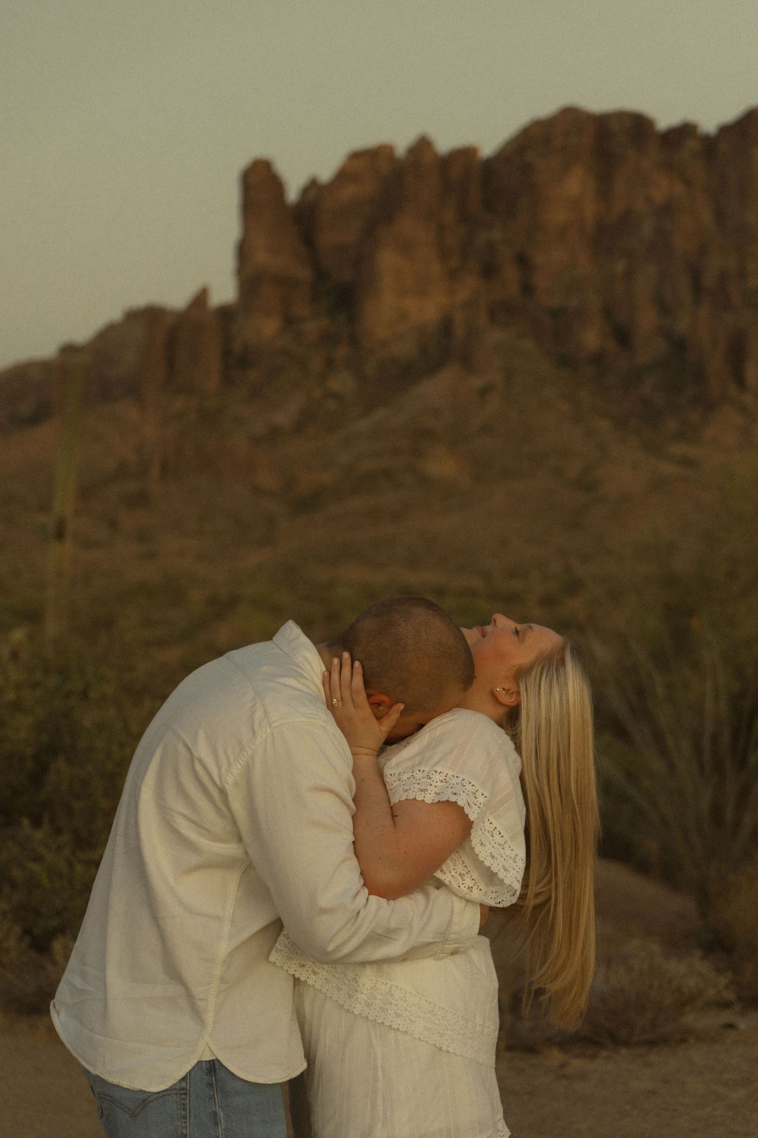 Couple kissing in white outfits at golden hour with Superstition Mountains and saguaro cacti in the Arizona desert background.