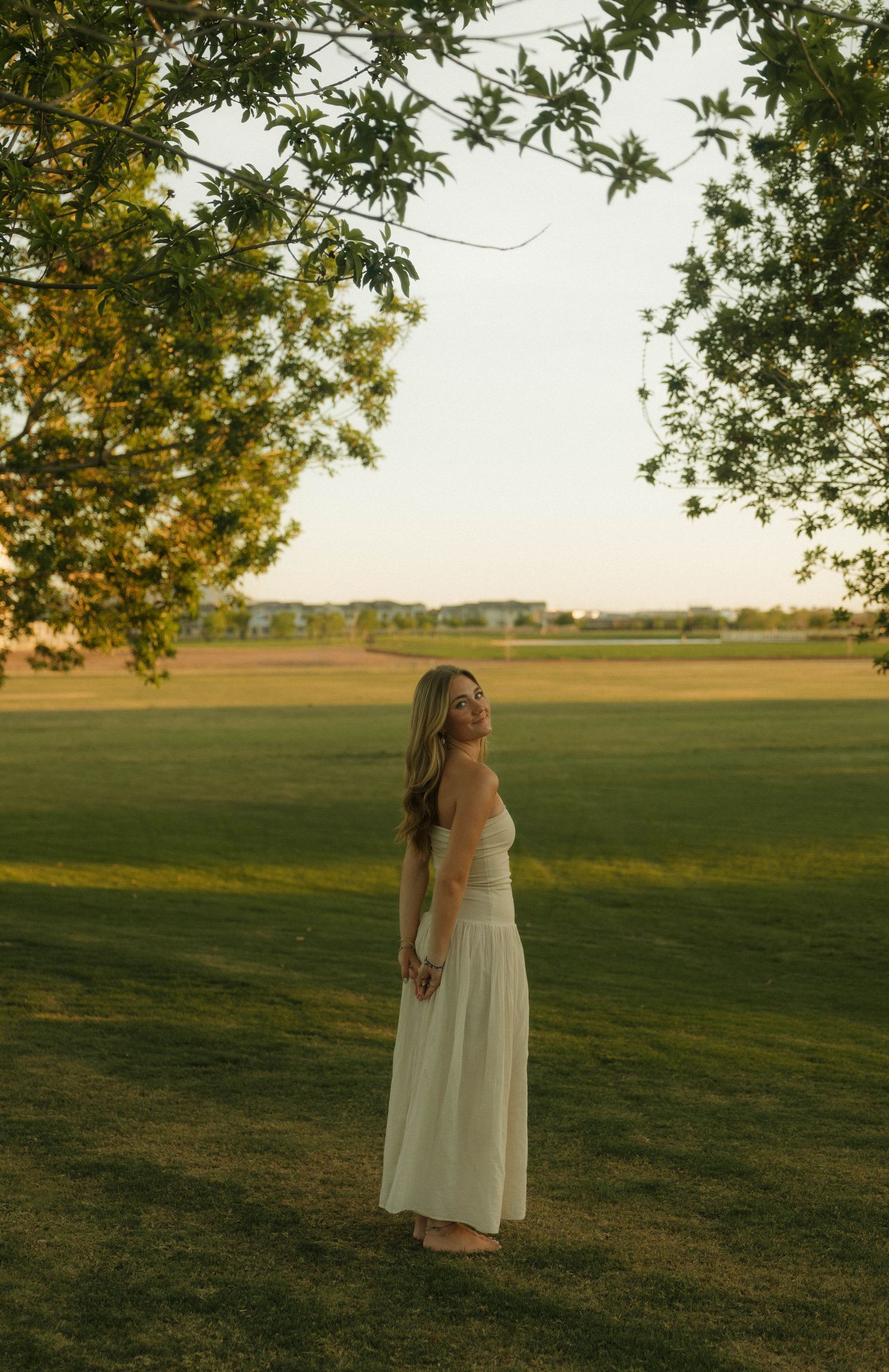 Woman in white maxi dress standing on green grass field under tree branches at golden hour sunset.