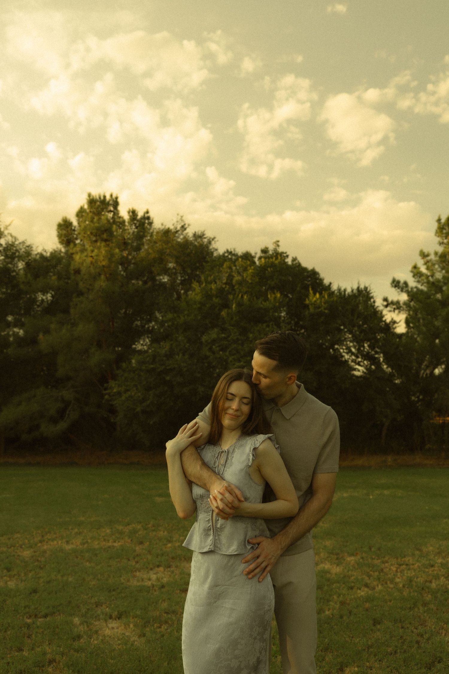 A couple embraces tenderly in a lush green park at golden hour, the man kissing the woman's head as she smiles softly.
