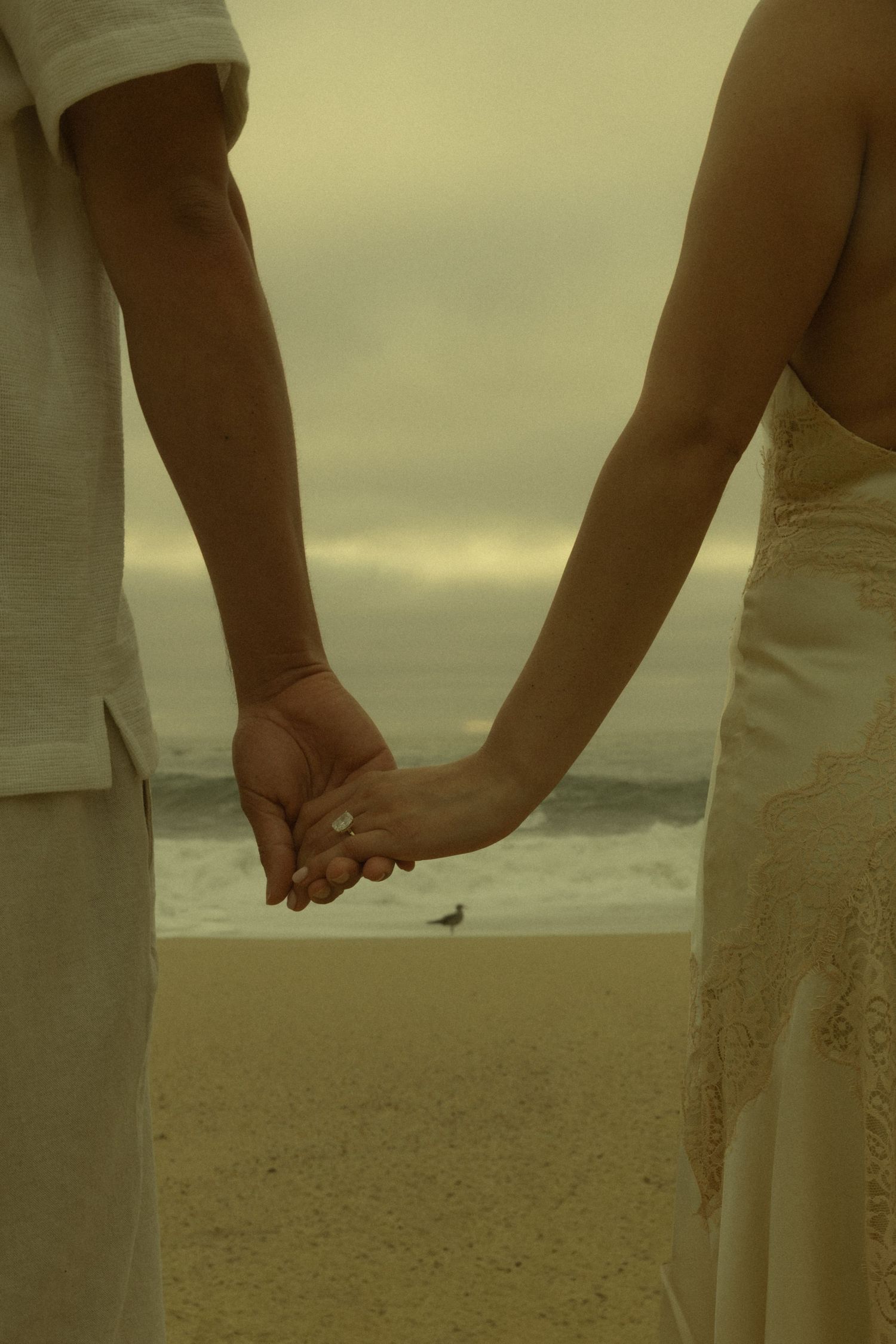 A couple holds hands on a sandy beach with ocean waves and cloudy sky in the background.