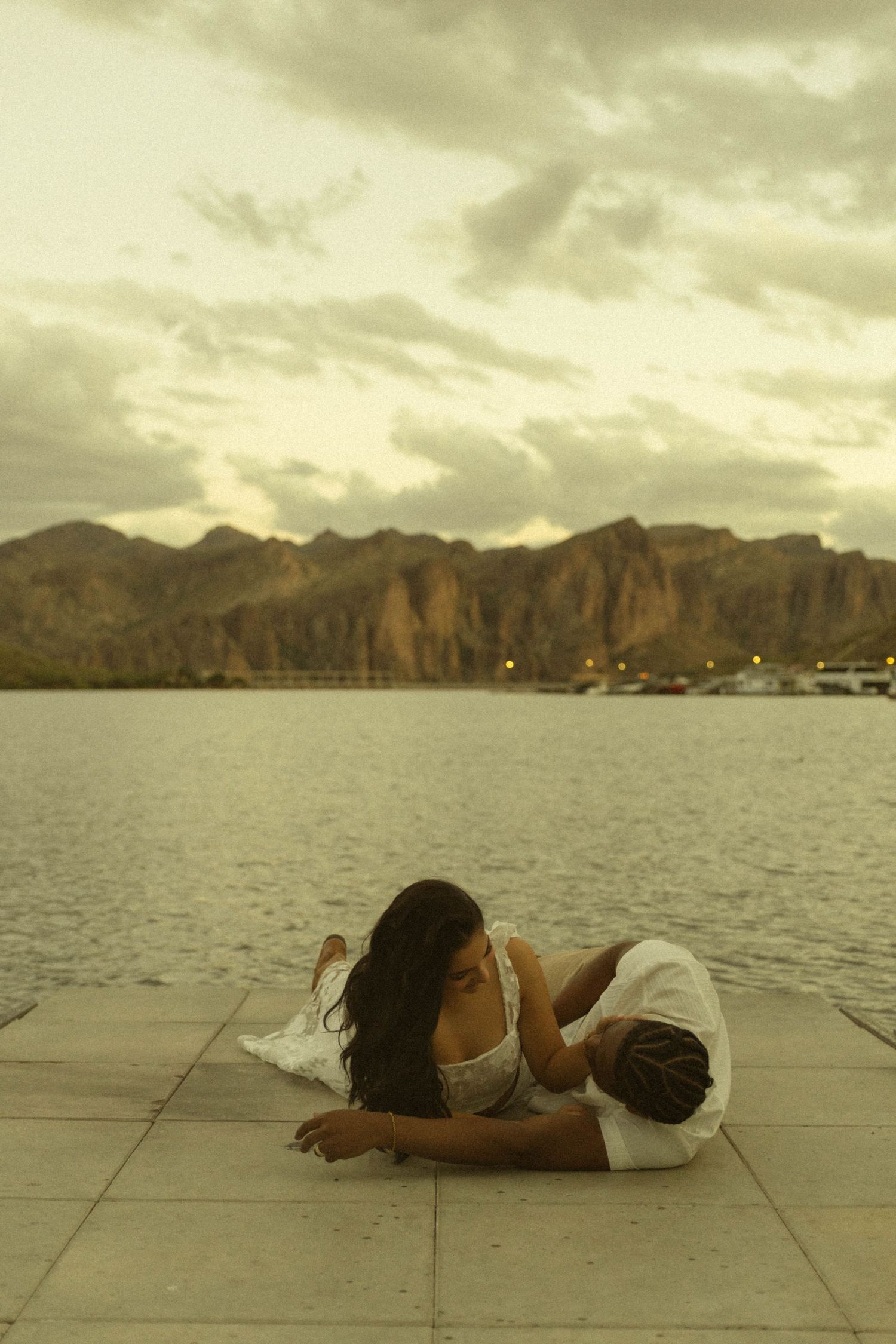 Romantic couple lying on waterfront tiles at sunset with mountain lake and cloudy sky in the background.