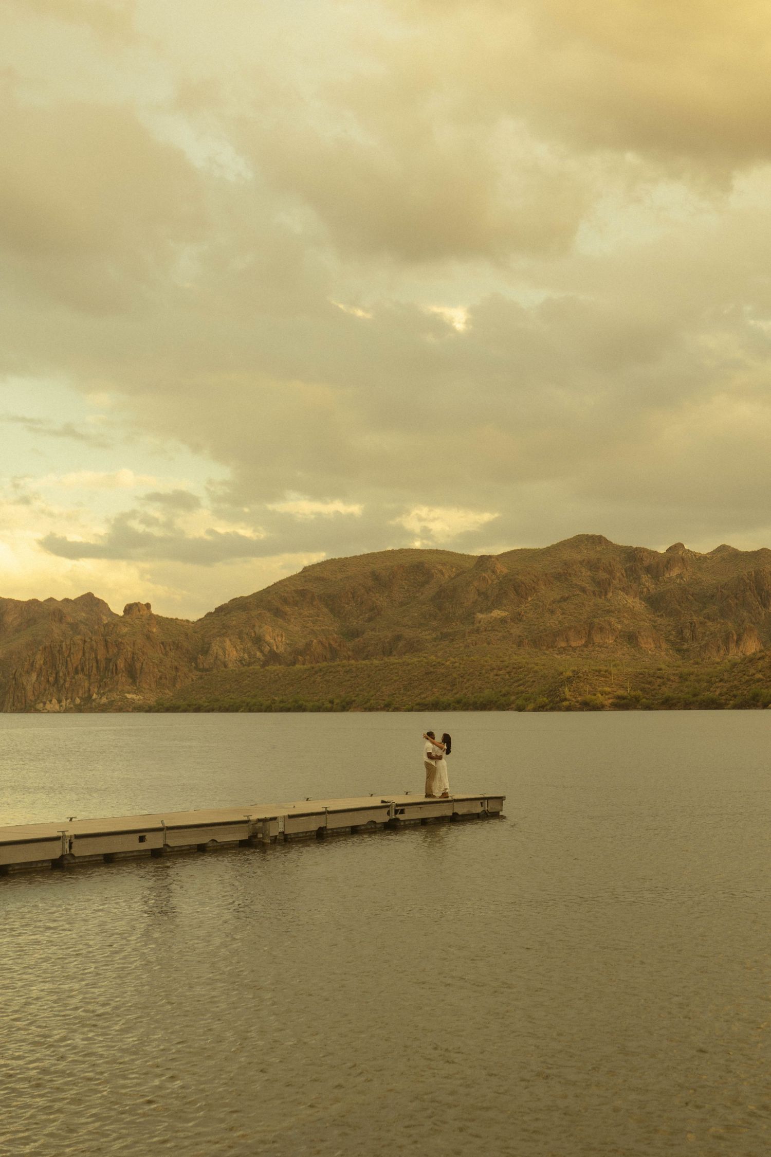 Couple embracing on a wooden dock at sunset over a calm lake with dramatic mountain backdrop.