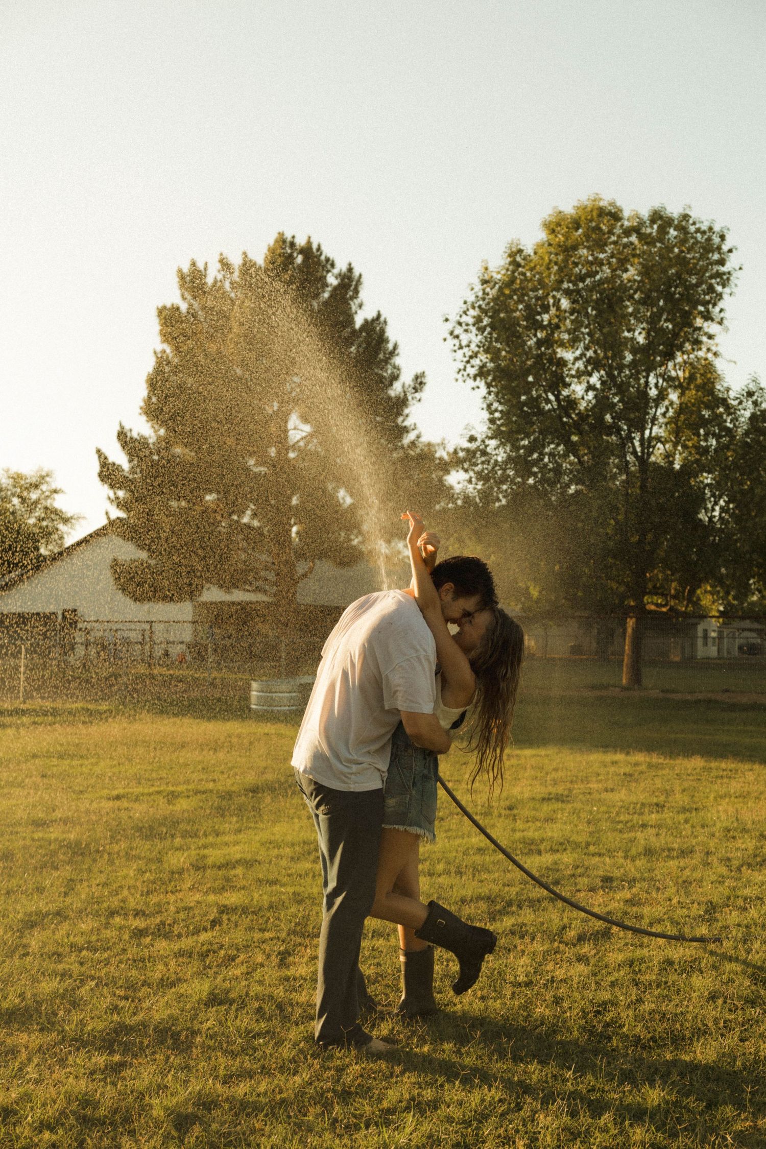 Couple embracing and kissing in a backyard with water spraying from a garden hose at golden hour.