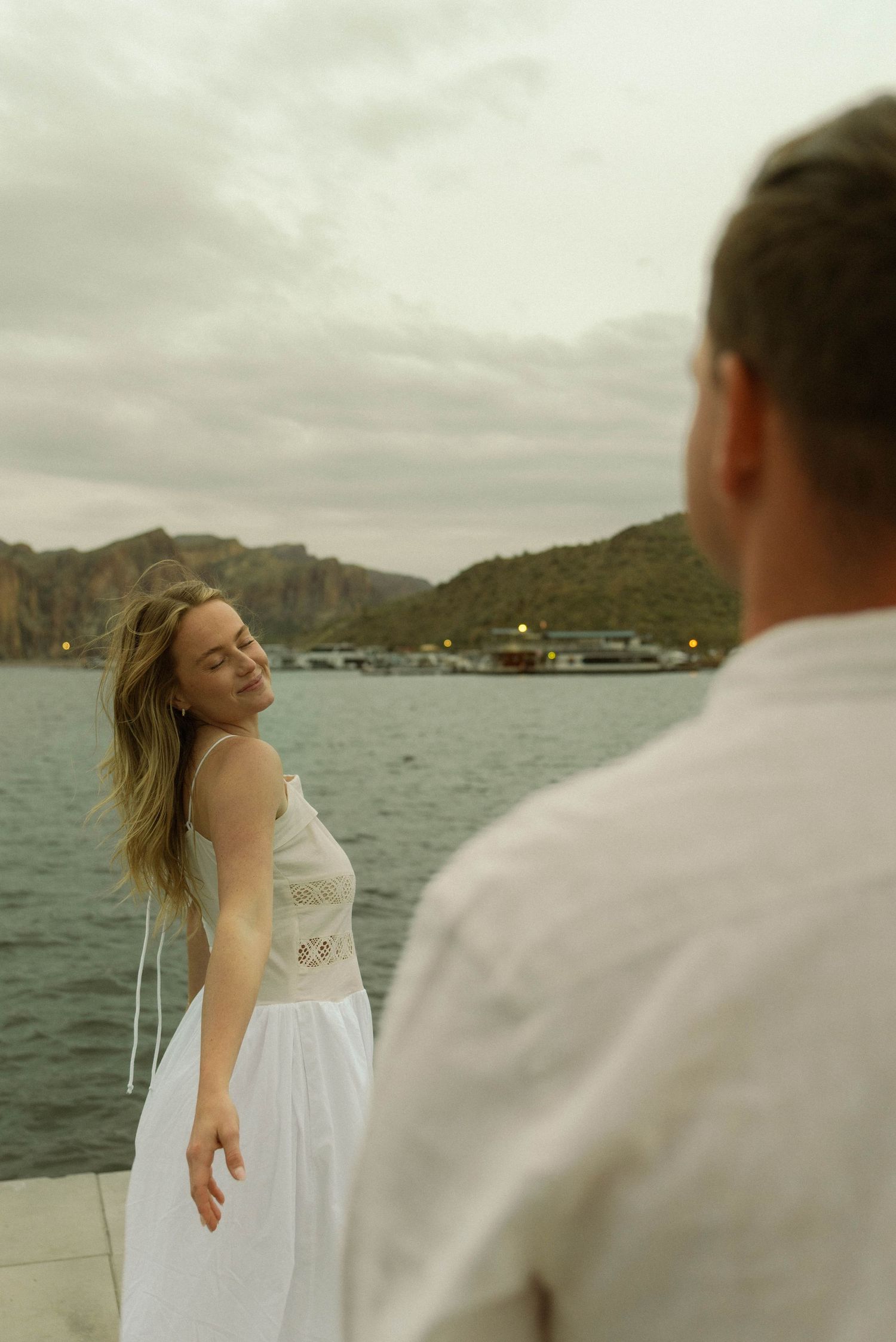 A couple shares a carefree moment on a boat with mountains and cloudy skies in the background.