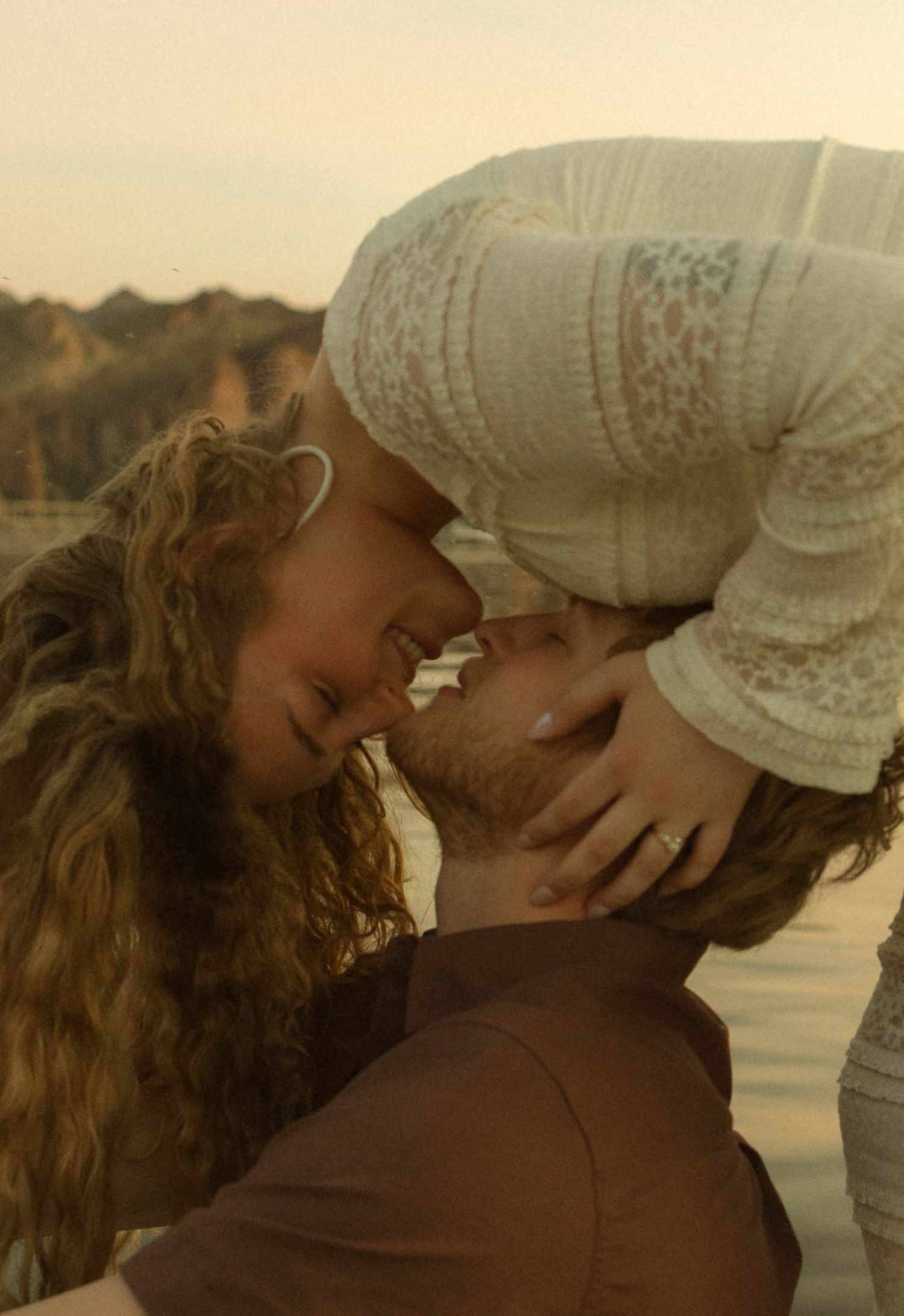 A romantic couple embraces and kisses at sunset by a tranquil lake with golden light illuminating their silhouettes.