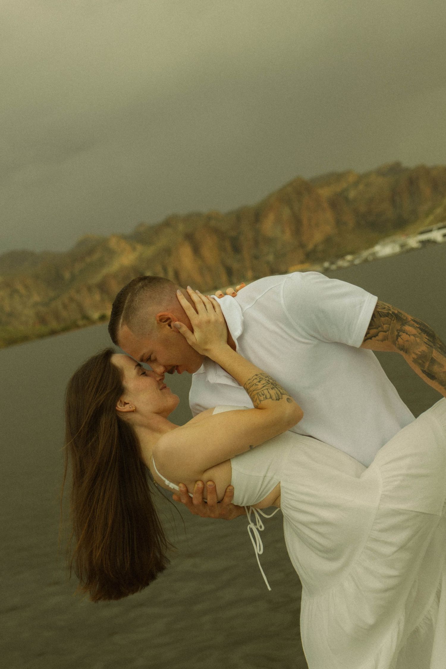 A romantic couple in white clothing share an intimate moment against a dramatic mountain backdrop near water.