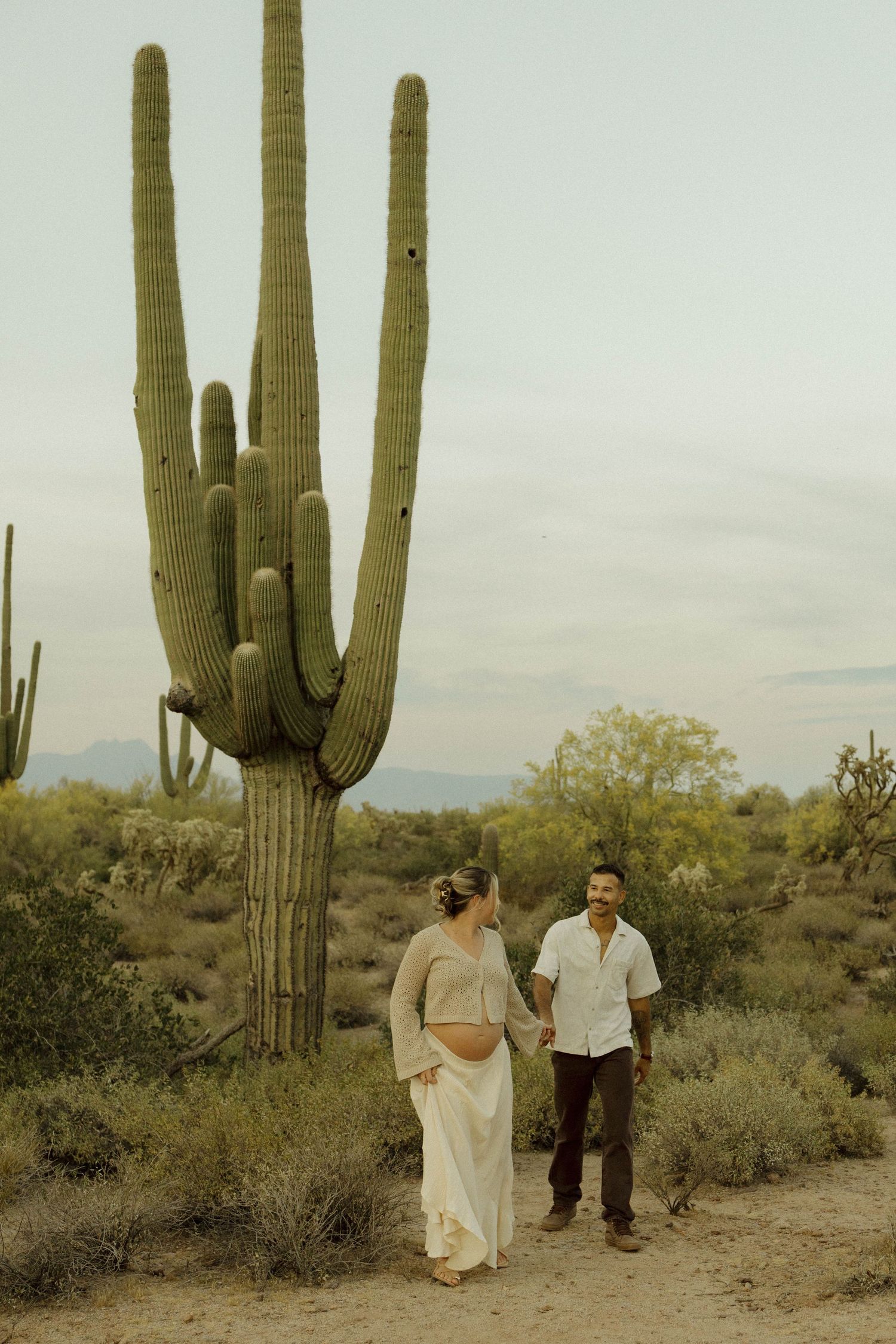 Expecting couple holding hands beside tall saguaro cactus in Arizona desert landscape.