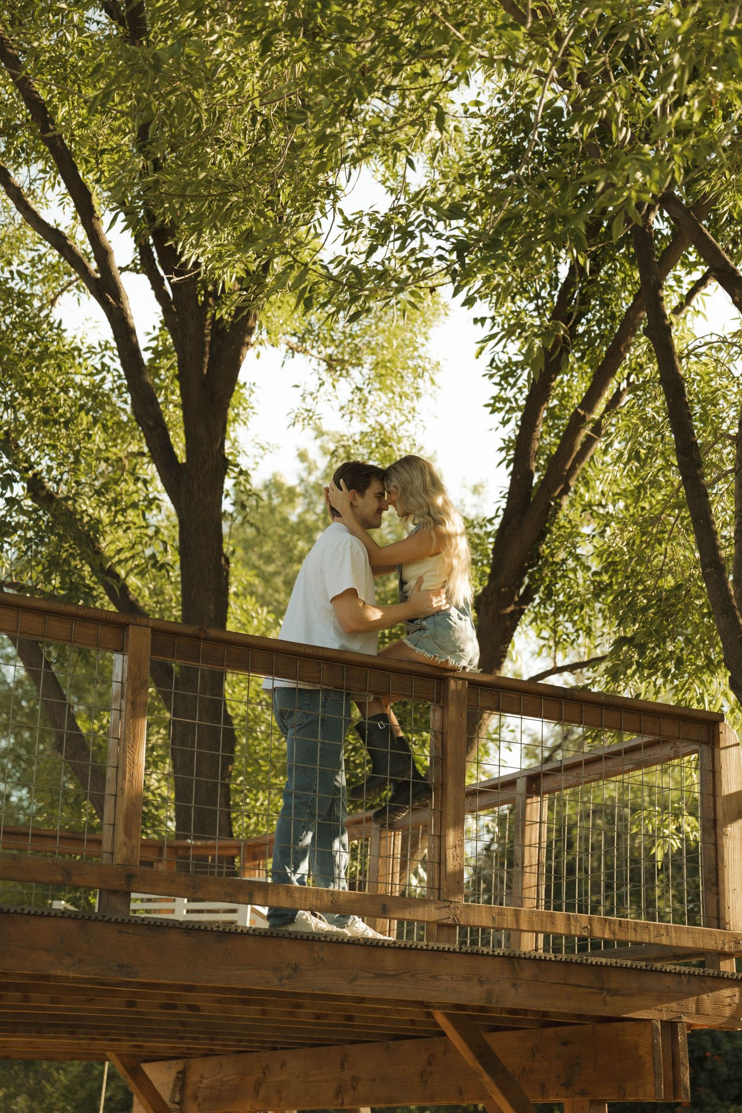 A romantic couple sharing a kiss on a wooden treehouse deck surrounded by lush green trees.