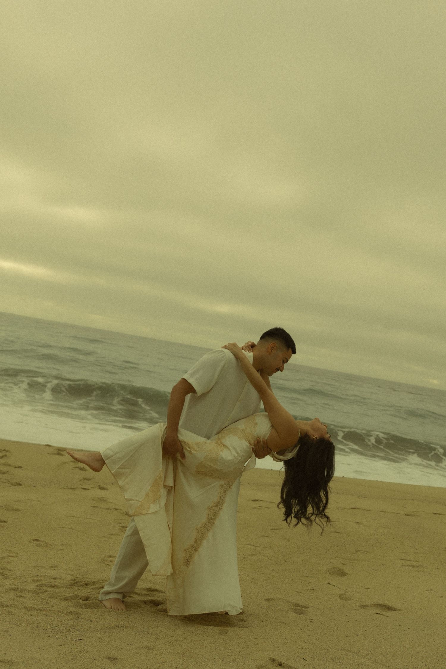 Couple dancing romantically on a sandy beach, man dipping woman in white dress, overcast sky and ocean waves in background.