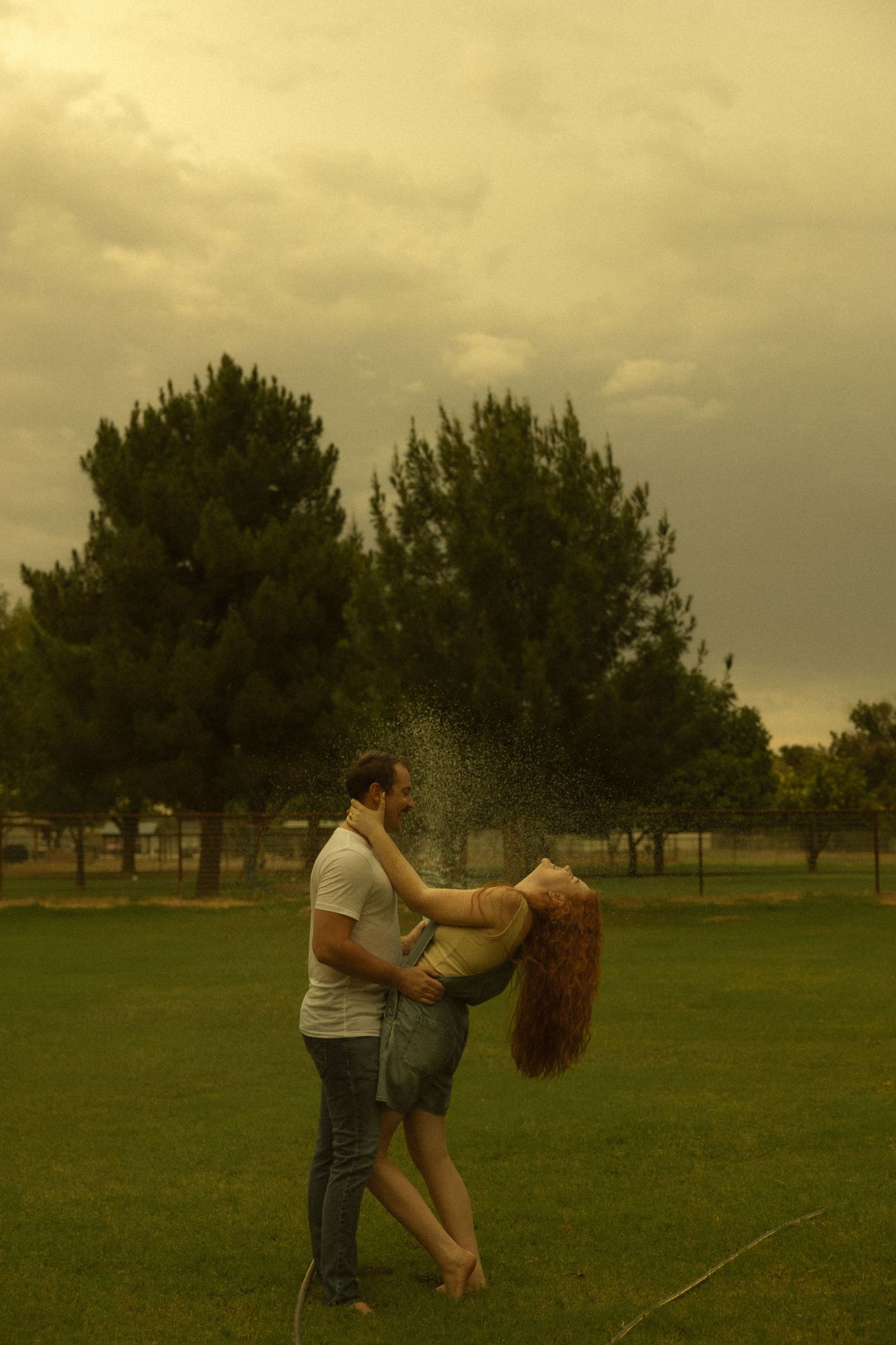 Couple embracing and dancing in a green park with water mist and moody cloudy sky behind them.