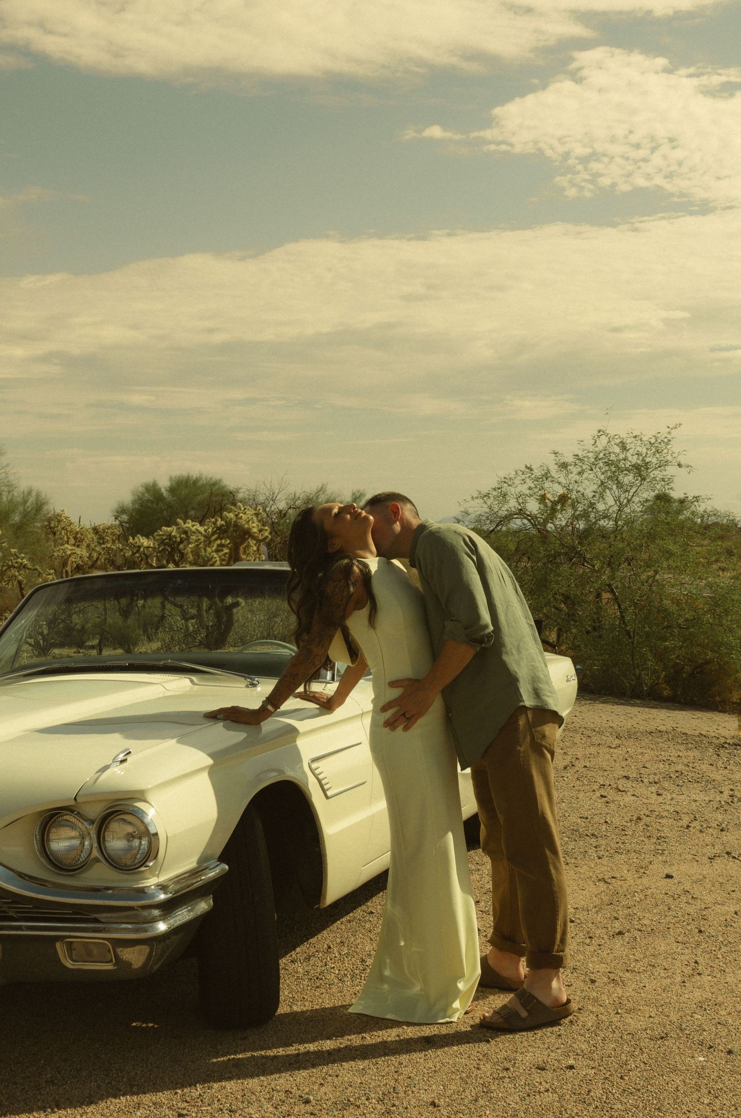 Couple kissing beside a vintage white convertible car in a desert landscape on their wedding day.