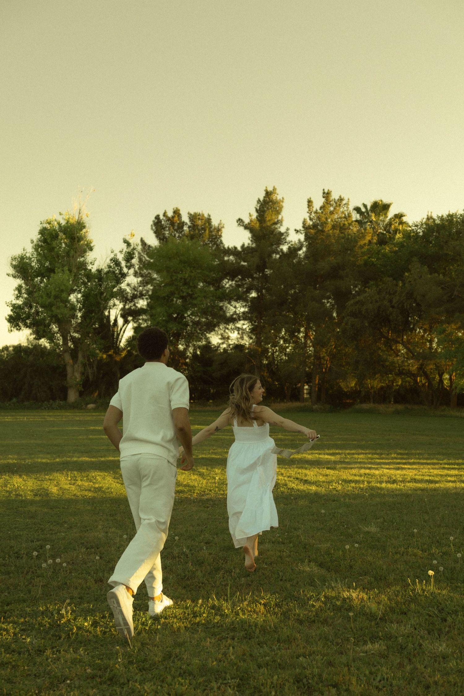 Couple holding hands running through a green park at sunset, both dressed in white outfits.