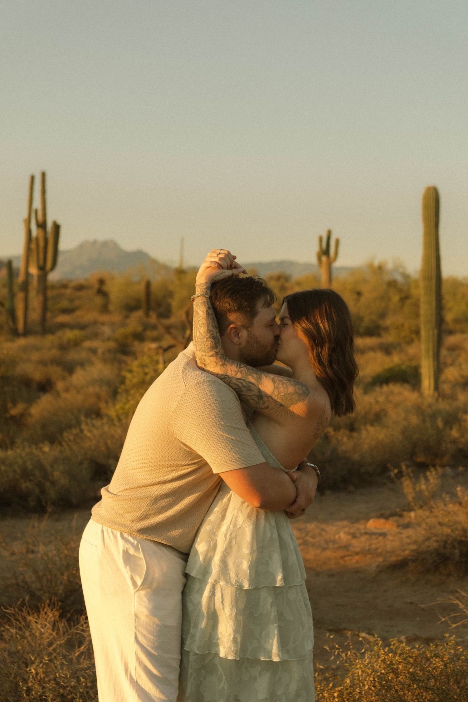 Couple sharing a romantic kiss at golden hour in the Arizona desert with saguaro cacti in the background.