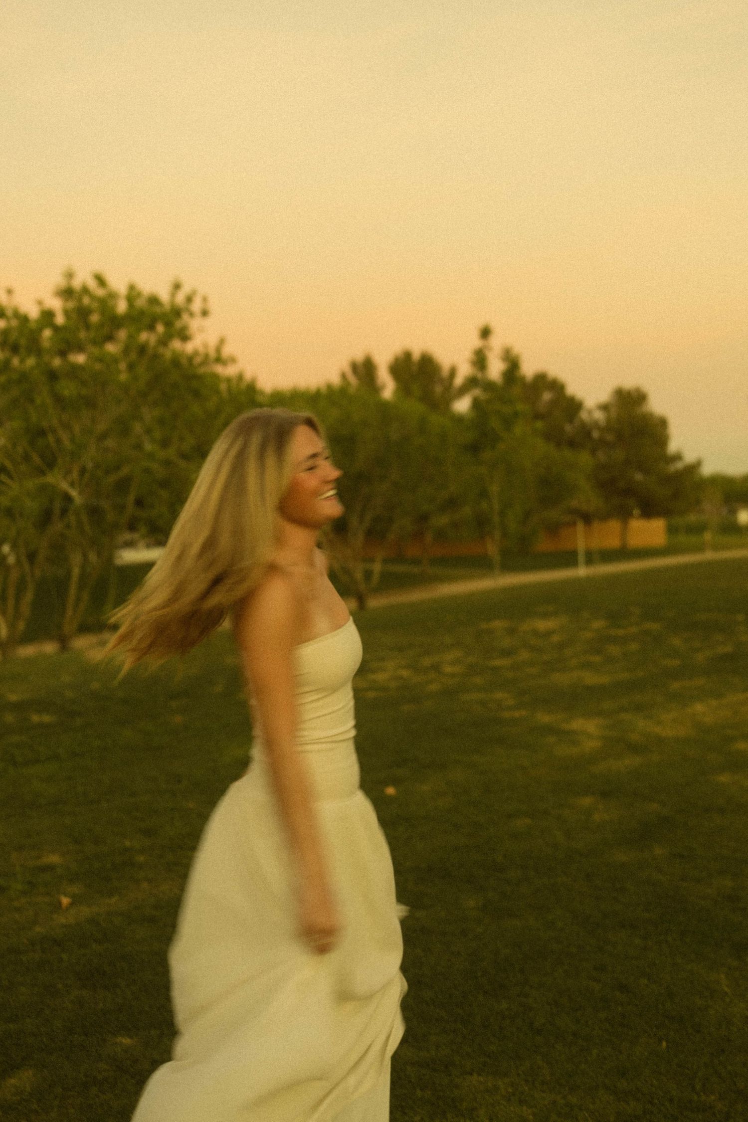 Smiling blonde woman in white dress twirling on green grass at golden hour sunset.