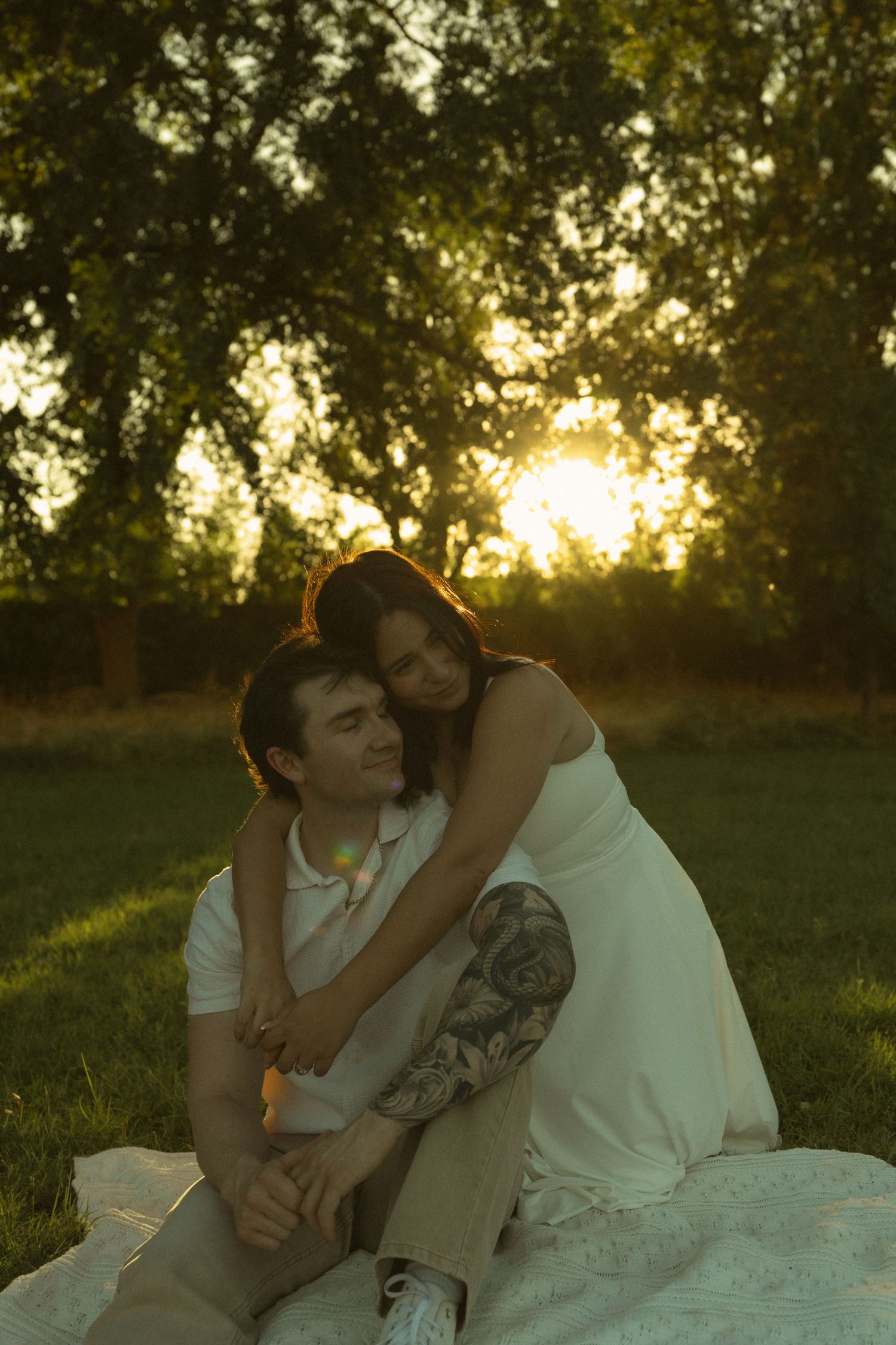 Romantic couple sitting on grass at sunset, woman in white dress with tattoos tenderly embracing man from behind.
