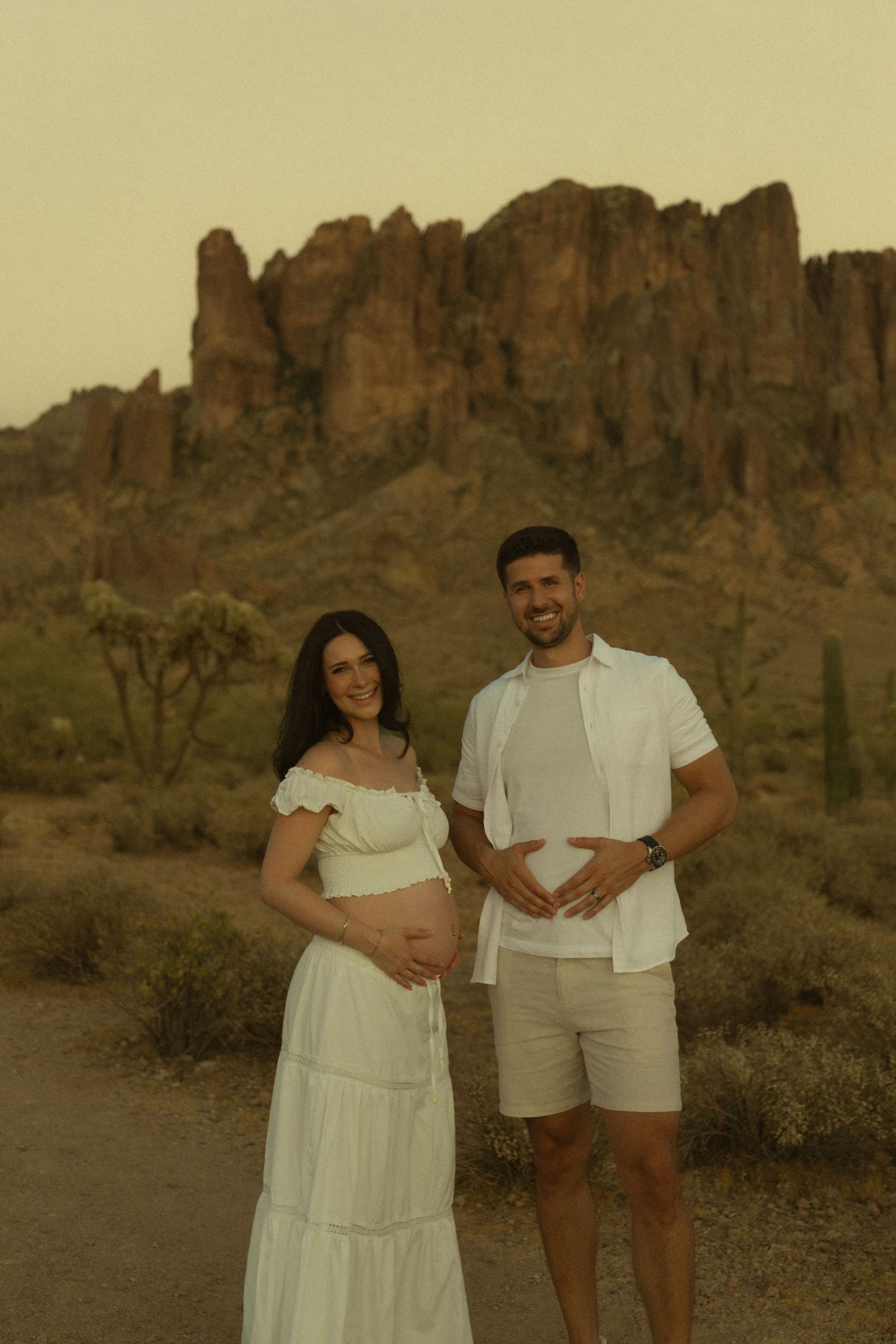 Pregnant couple in white posing at golden hour with Superstition Mountains in Arizona desert.