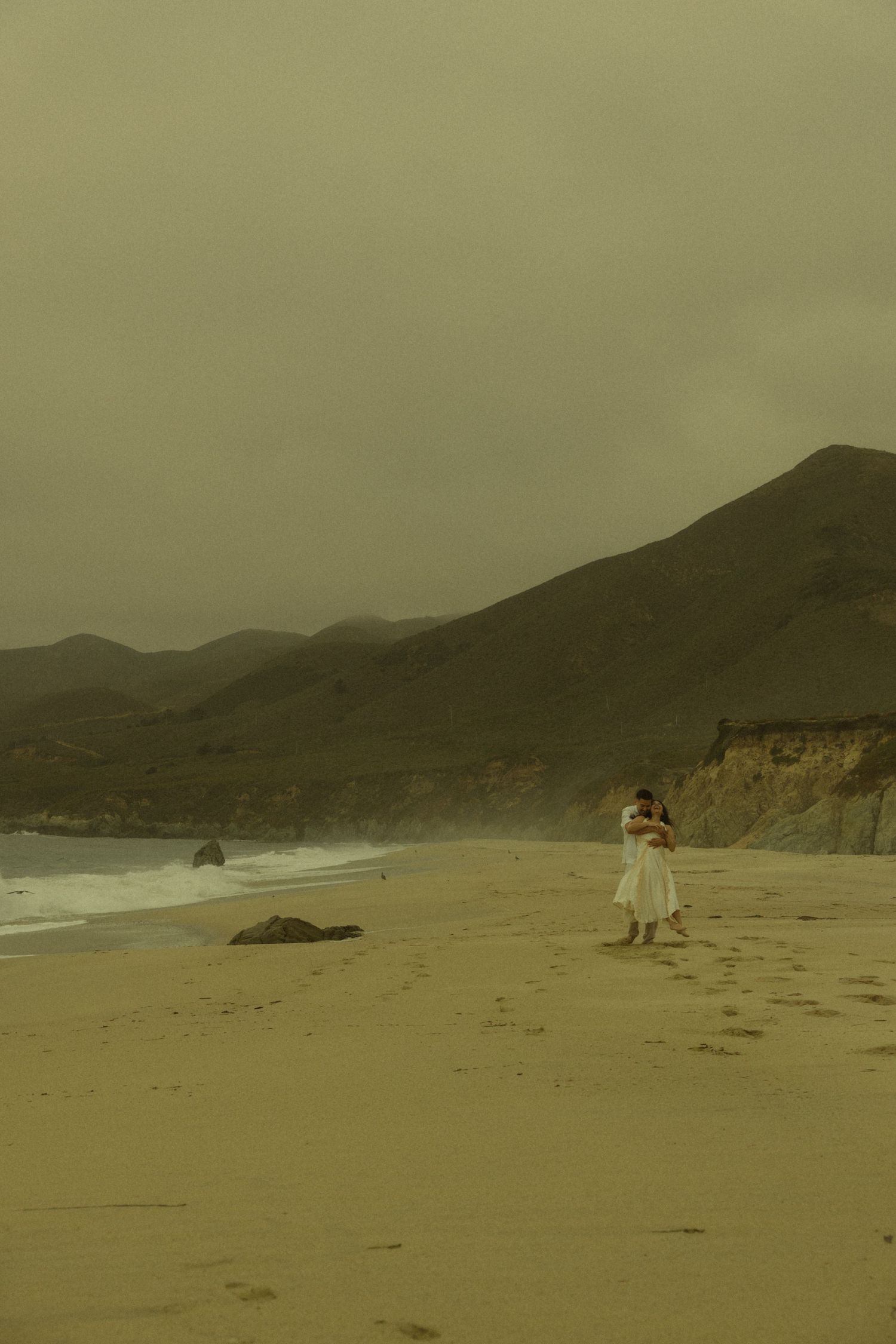 Couple embracing on a misty sandy beach with dramatic coastal cliffs and moody overcast skies in the background.
