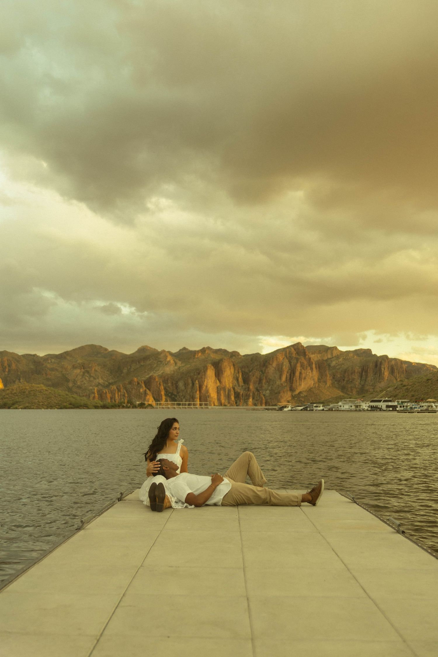 Couple relaxing on a dock at sunset with dramatic mountain backdrop and cloudy sky over calm lake waters.