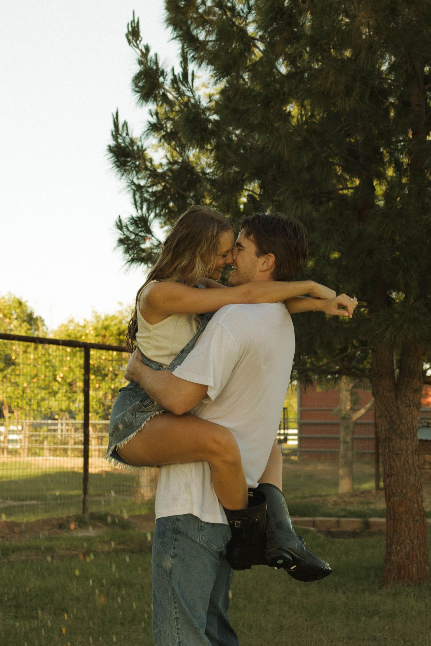 Couple sharing a romantic embrace outdoors near a pine tree at golden hour, man lifting woman in jeans and boots.