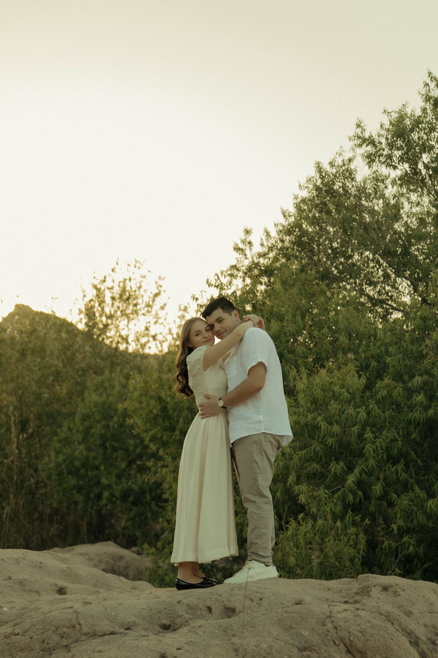 Couple embracing on rocky terrain with lush green trees at golden hour sunset.
