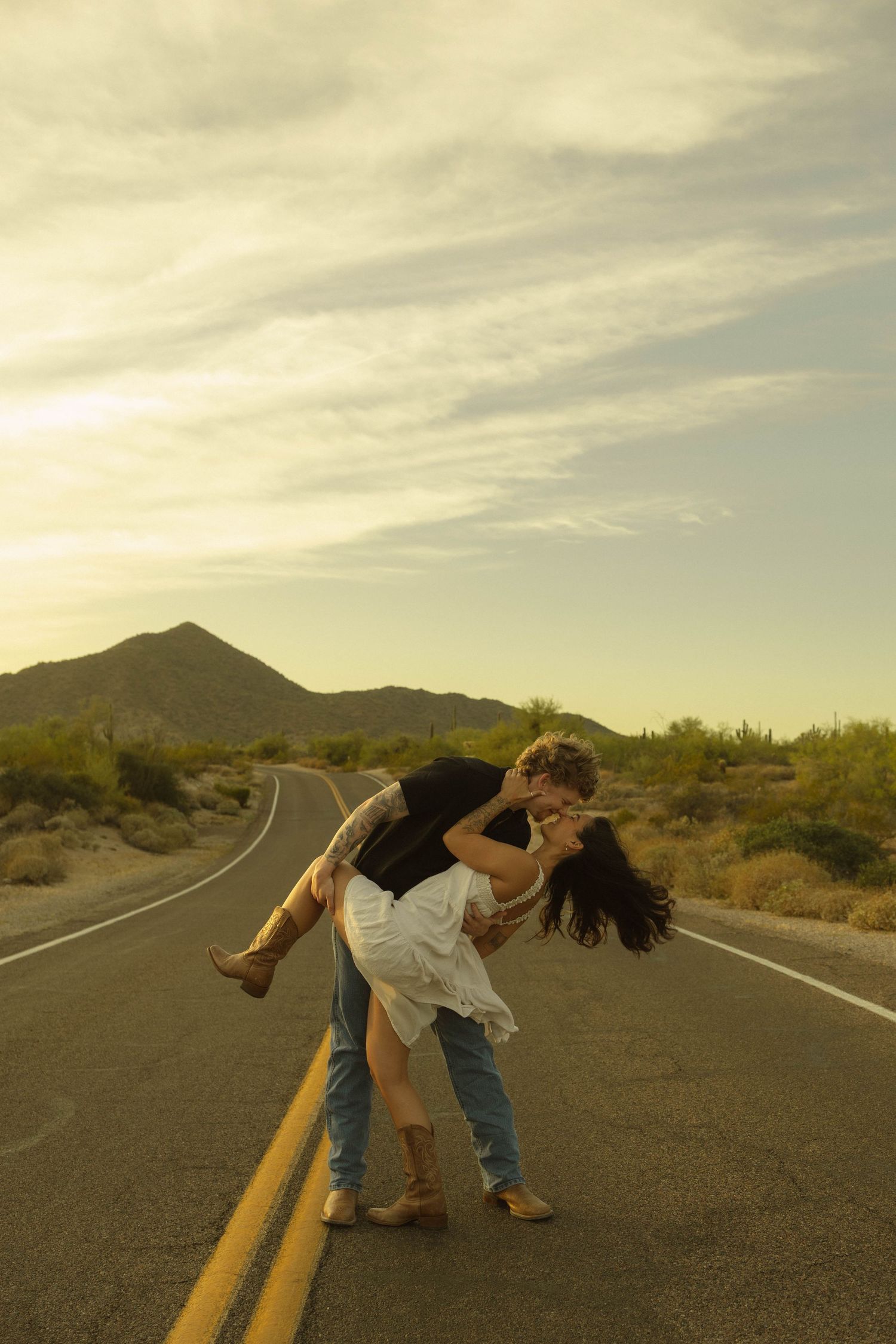 Couple sharing a passionate dip kiss on a desert road at sunset with scenic mountain backdrop.