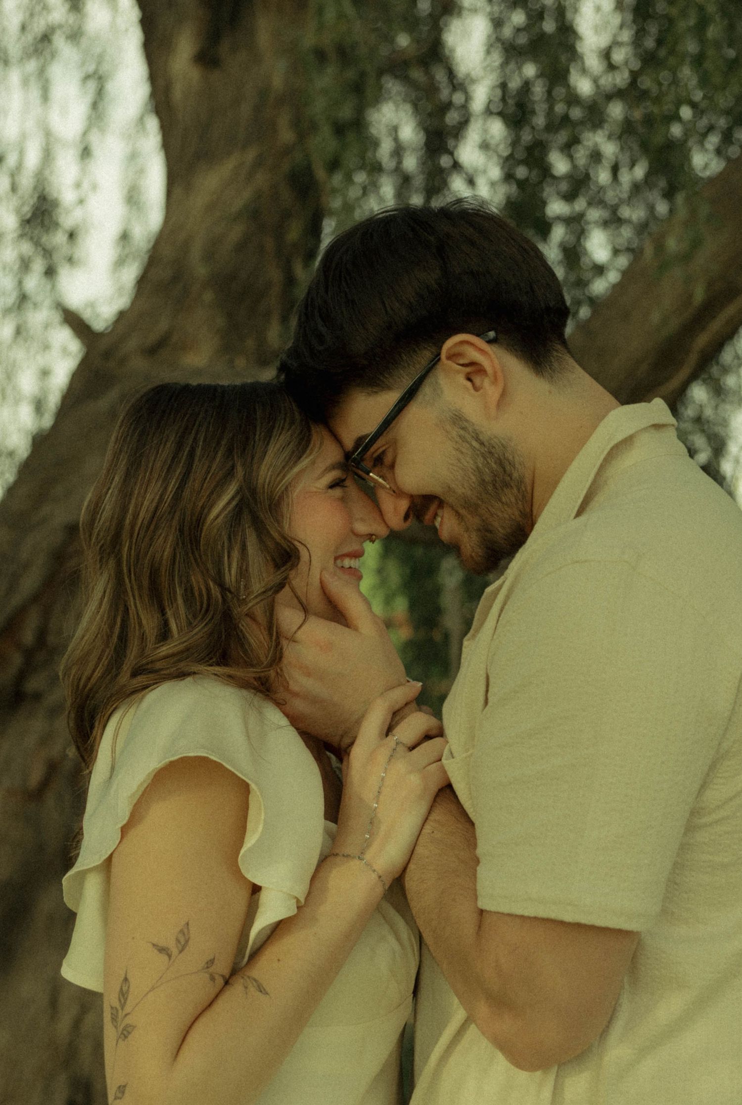 Romantic couple smiling and touching foreheads outdoors near a tree, woman with floral tattoo wearing white dress.
