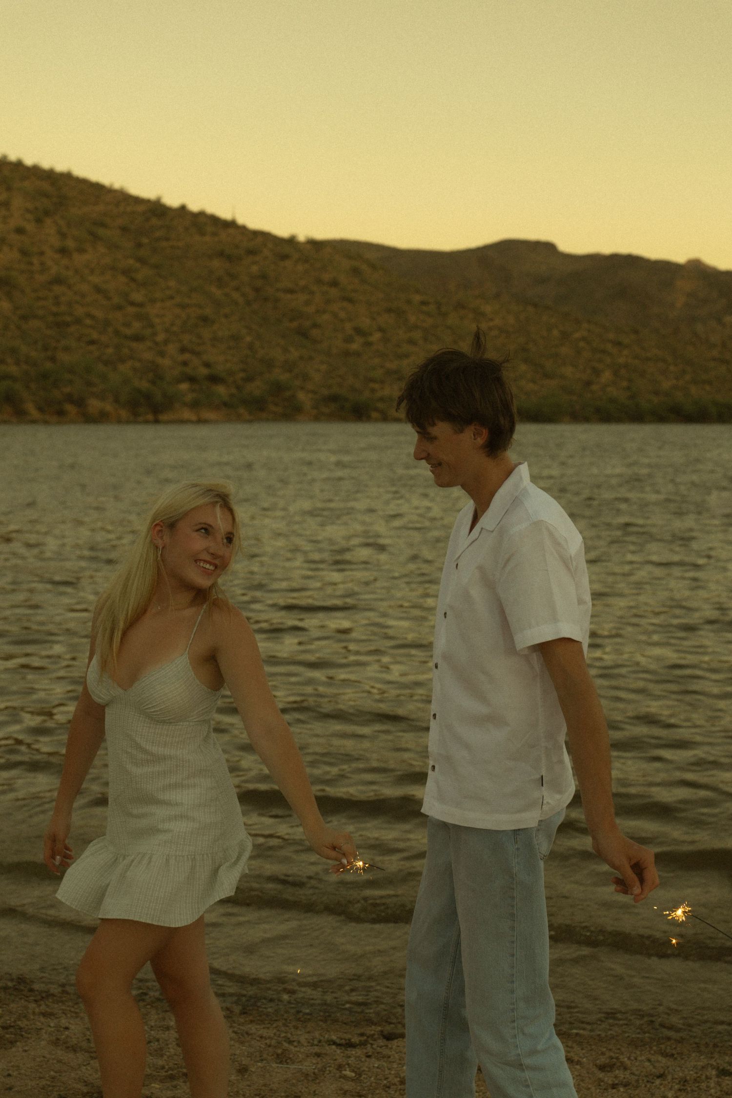 Young couple holding sparklers walking along a lakeside beach at sunset with green hills in the background.