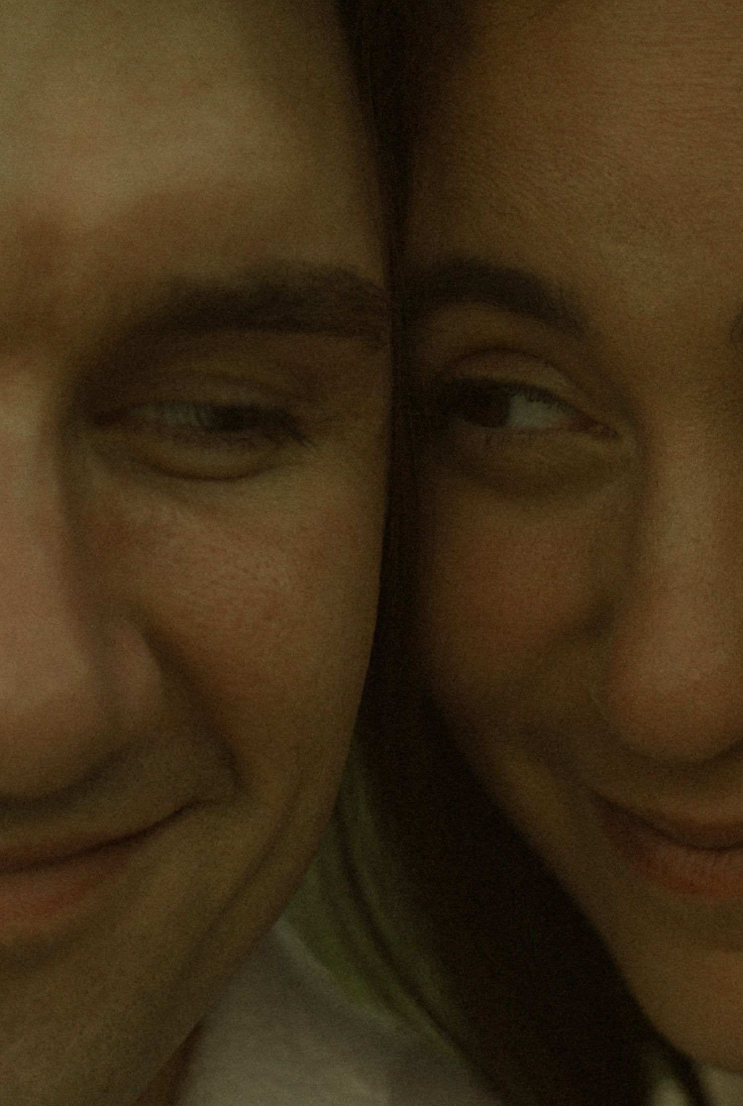 Close-up portrait of two smiling people with faces pressed together, captured in warm, dim lighting.