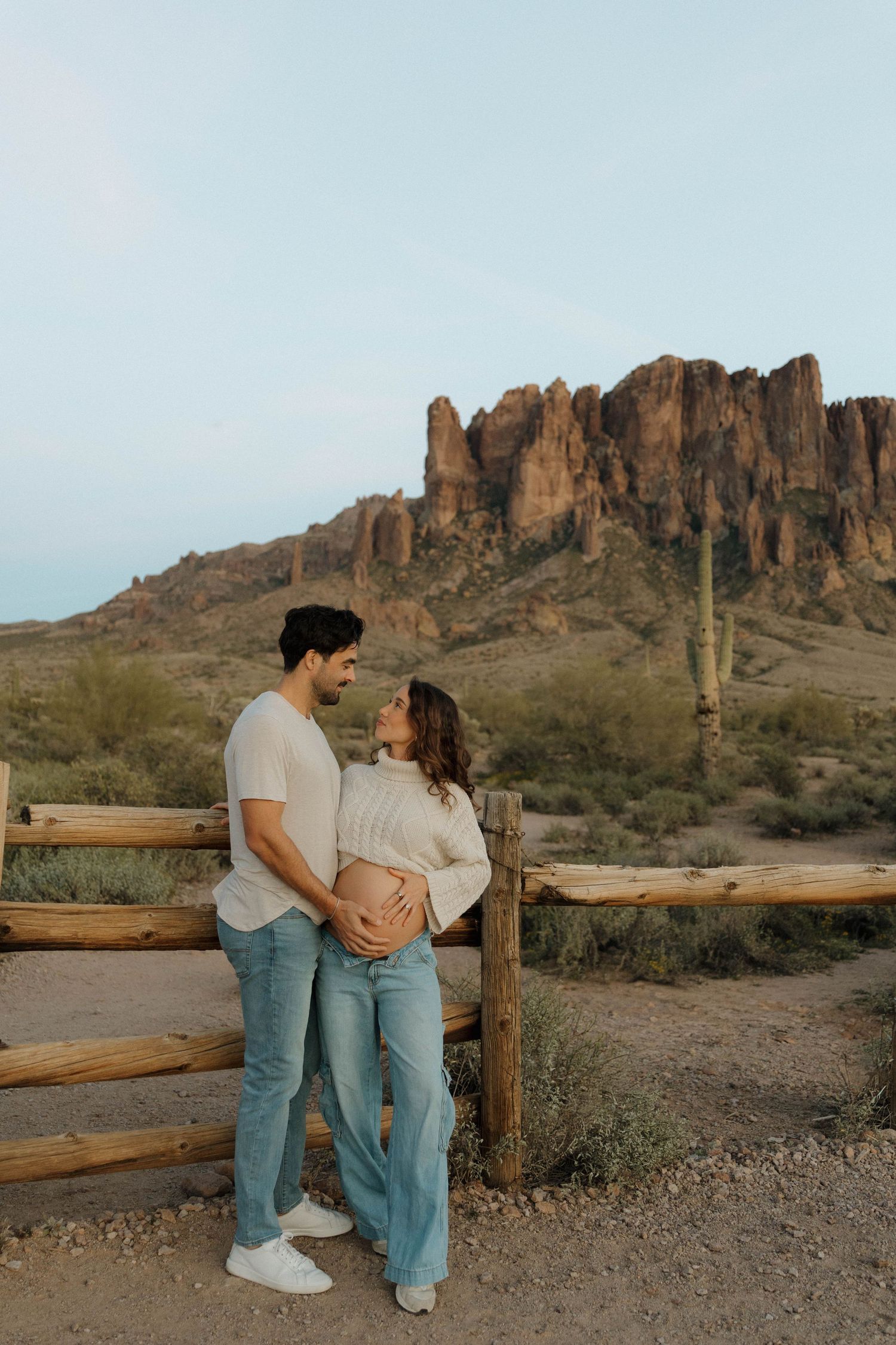 Expecting couple shares tender moment by wooden fence with Superstition Mountains in background.