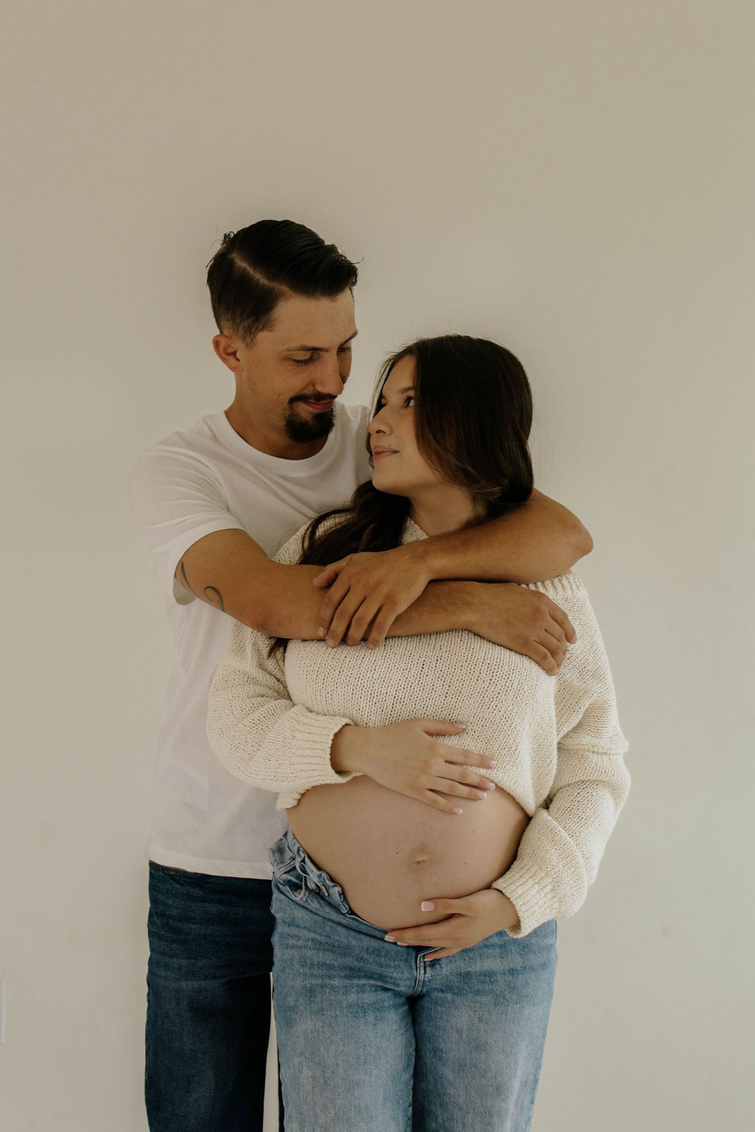 A couple embraces tenderly while wearing casual clothing against a neutral wall background.