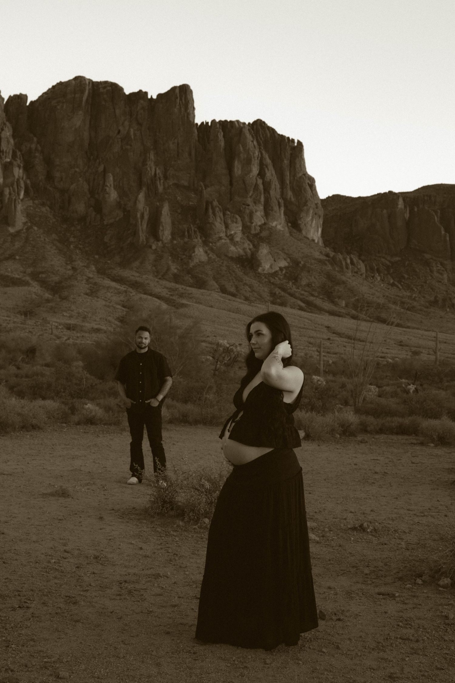 A couple in dark clothing poses playfully in front of desert cliffs in black and white.