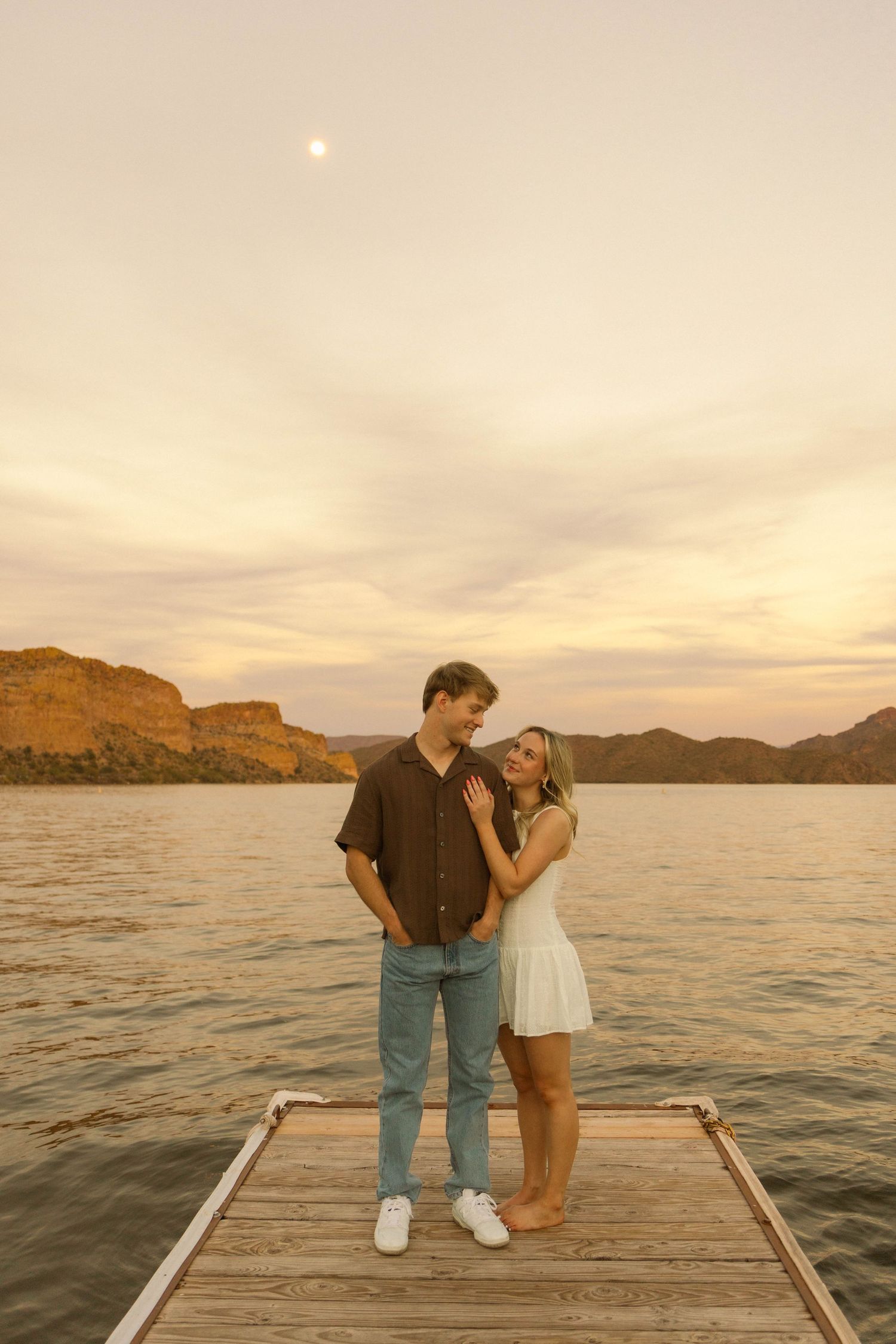 Couple stands together on wooden dock overlooking mountain lake at sunset in warm lighting.