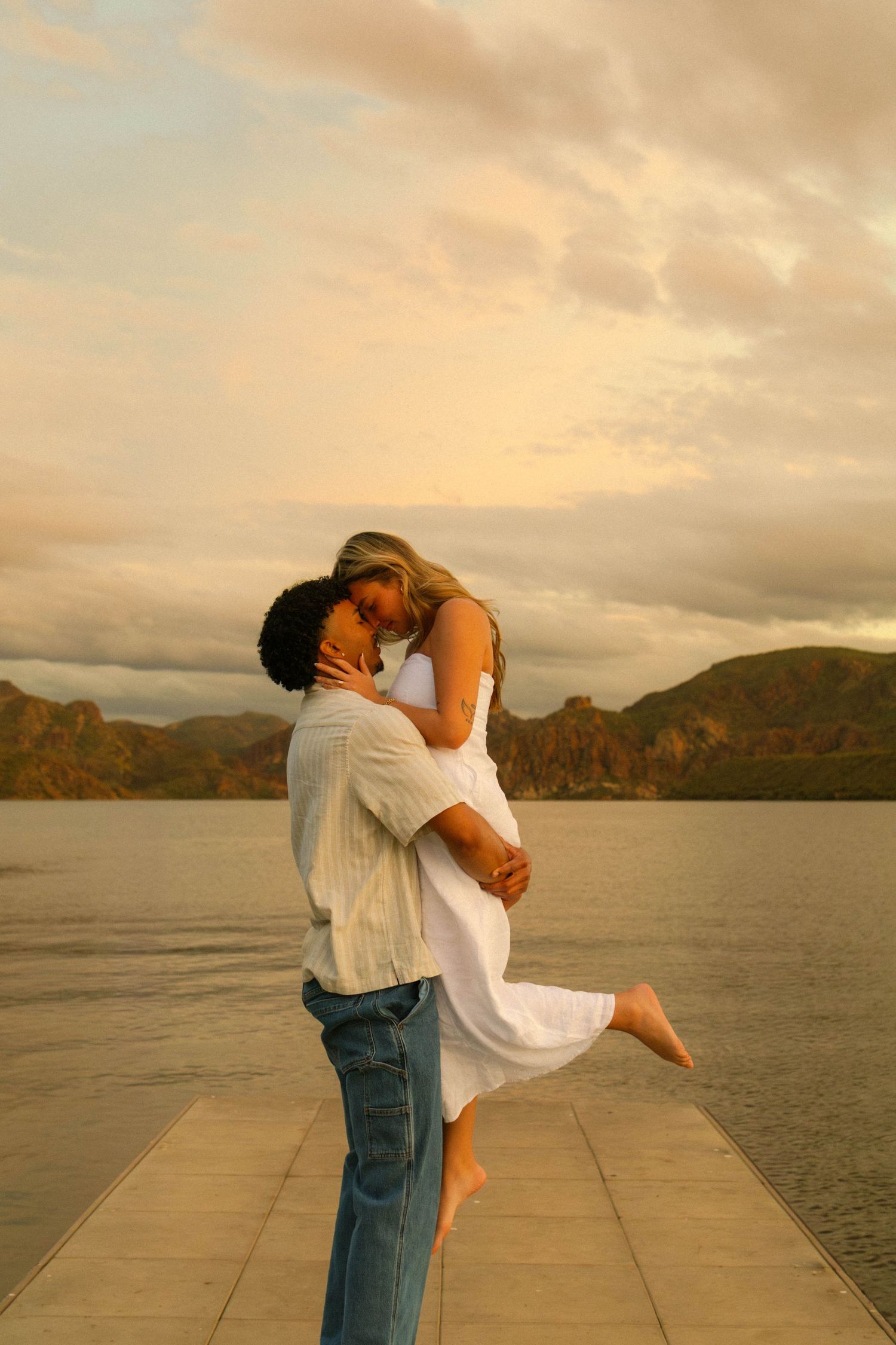 A romantic couple embraces on a dock by a lake at sunset with mountains in the background.