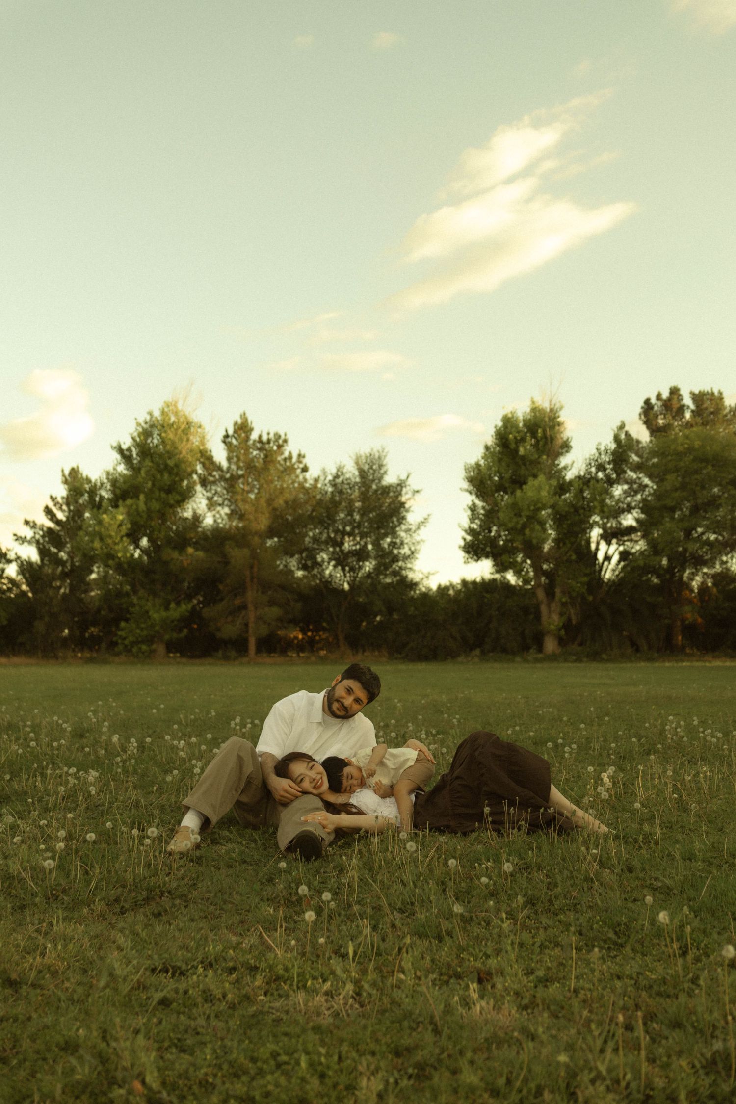 Family of three relaxing together on green grass in a sunlit park surrounded by tall trees at golden hour.