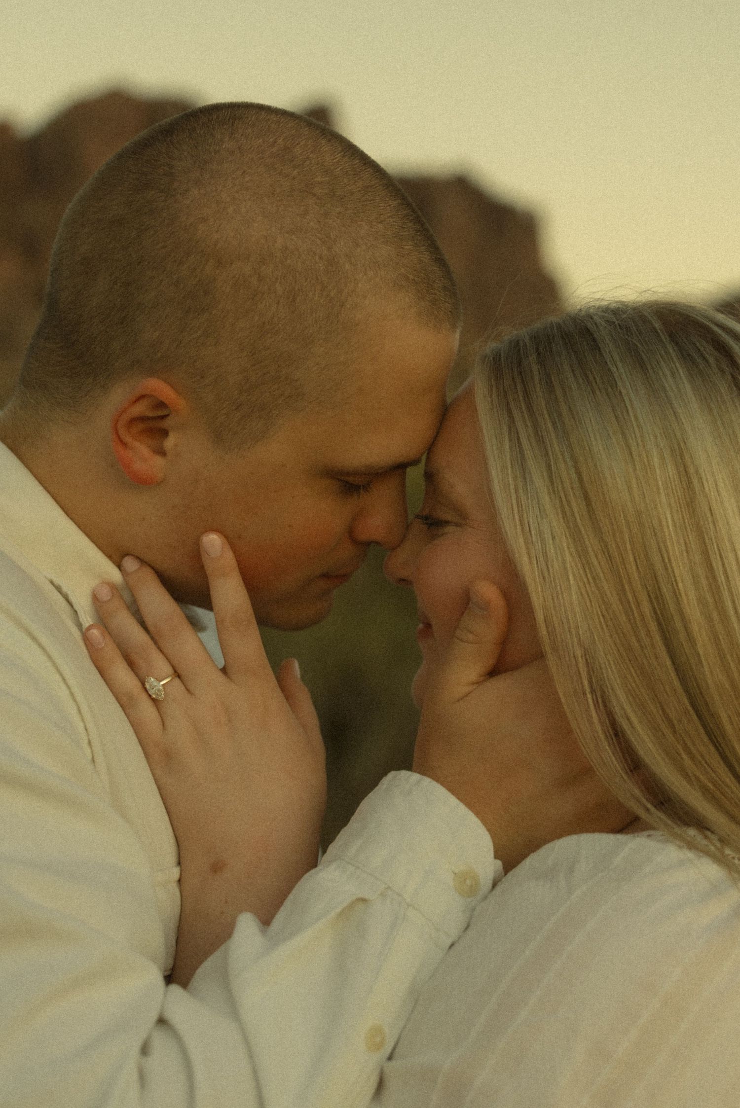 Romantic couple in white shirts touching foreheads tenderly, woman wearing engagement ring, soft golden light background.