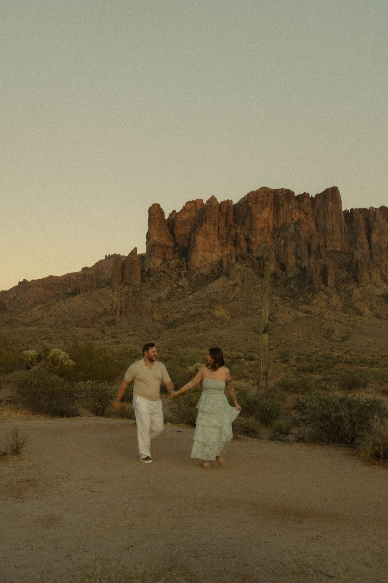 Couple strolling hand in hand through Arizona desert with rocky mountain backdrop at dusk.