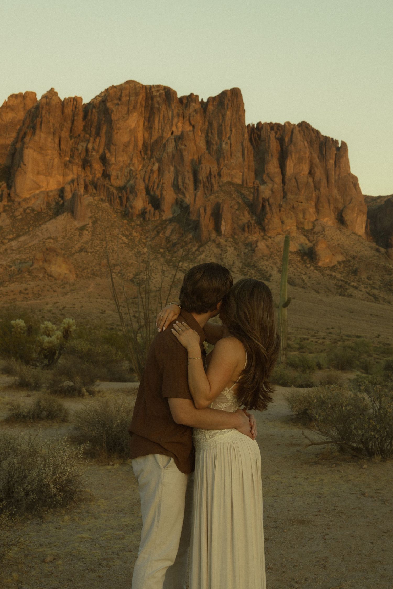 Couple embracing and kissing in Arizona desert with dramatic red rock Superstition Mountains at sunset.