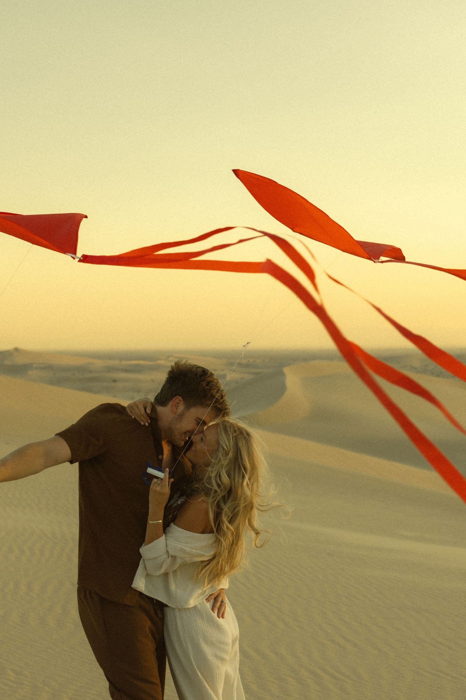 Couple kissing in desert dunes at sunset with red ribbons flying in the wind.