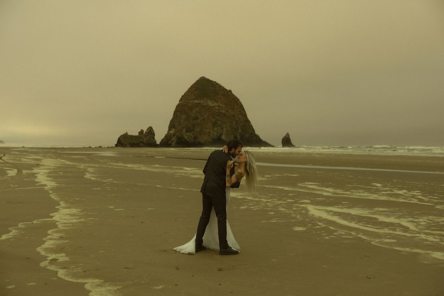 A couple shares a romantic kiss on a misty beach with Haystack Rock in the background at Cannon Beach, Oregon.
