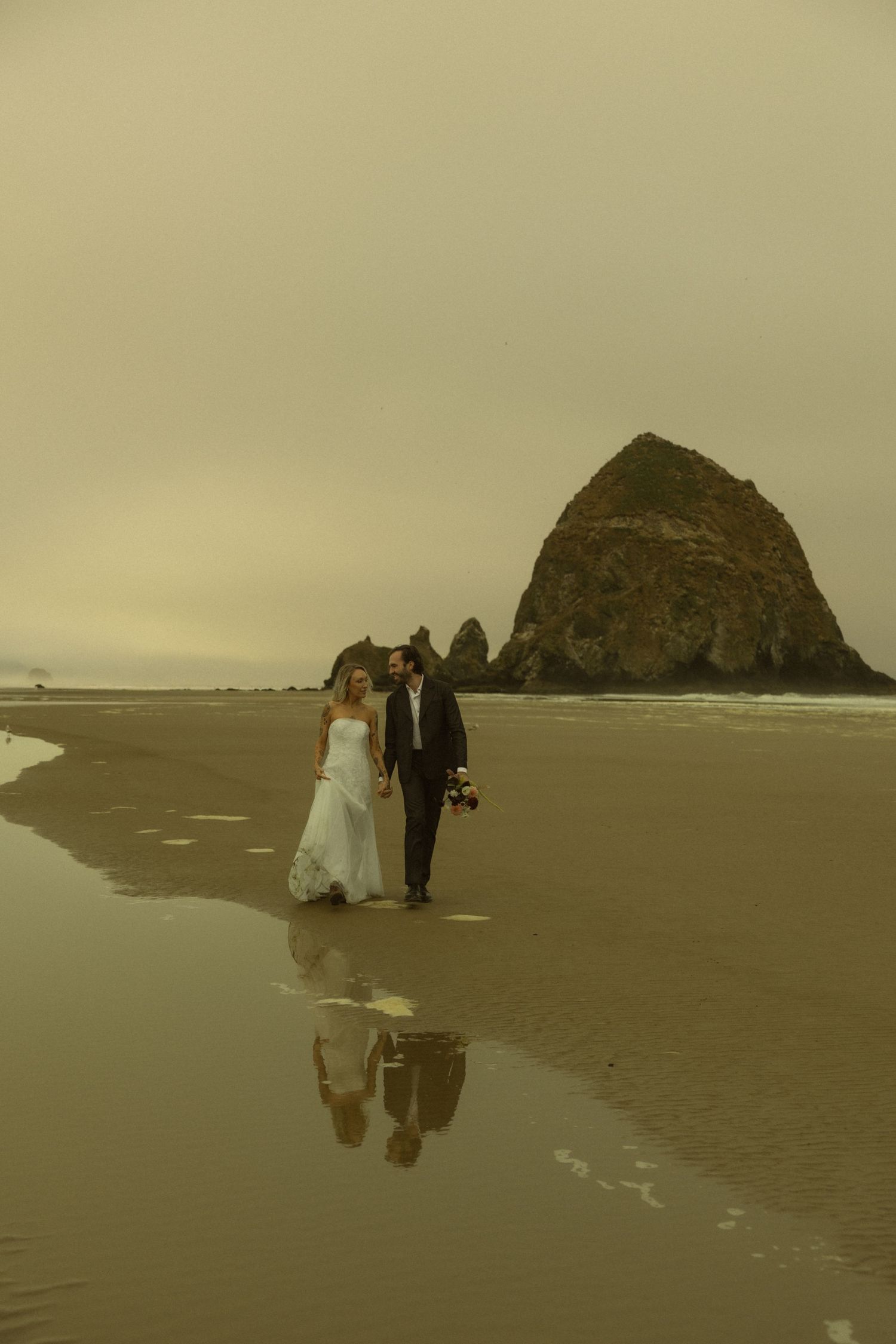 Bride and groom walk hand-in-hand on a reflective beach near Haystack Rock at Cannon Beach, Oregon.