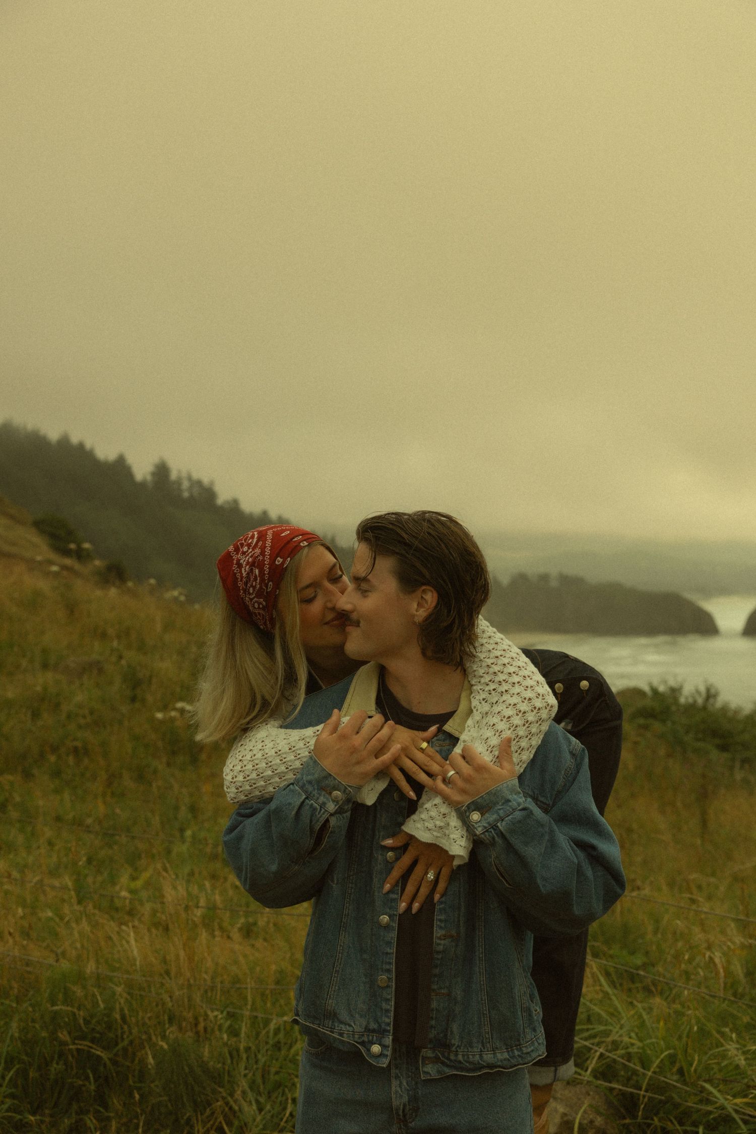 Couple sharing a romantic kiss on a misty coastal hillside, wearing denim and knit sweater, with ocean cliffs in background.