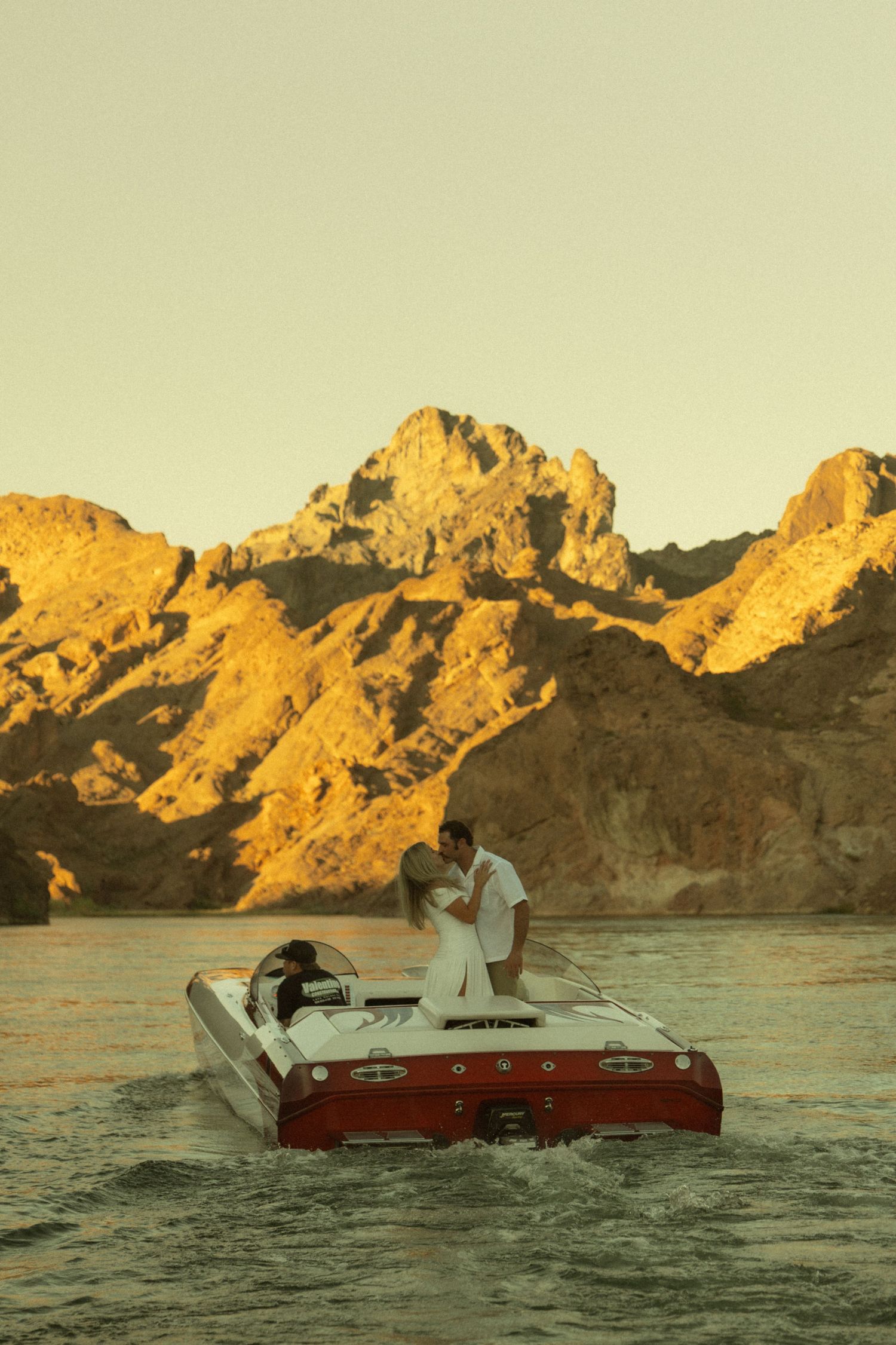 Couple kissing on a red and white speedboat at golden sunset with dramatic rocky mountains in background.