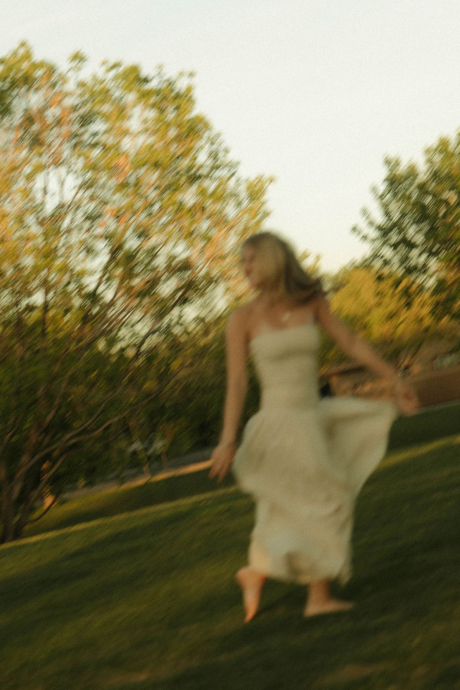 A blurred woman in a white dress runs barefoot across a lush green lawn surrounded by autumn trees at dusk.